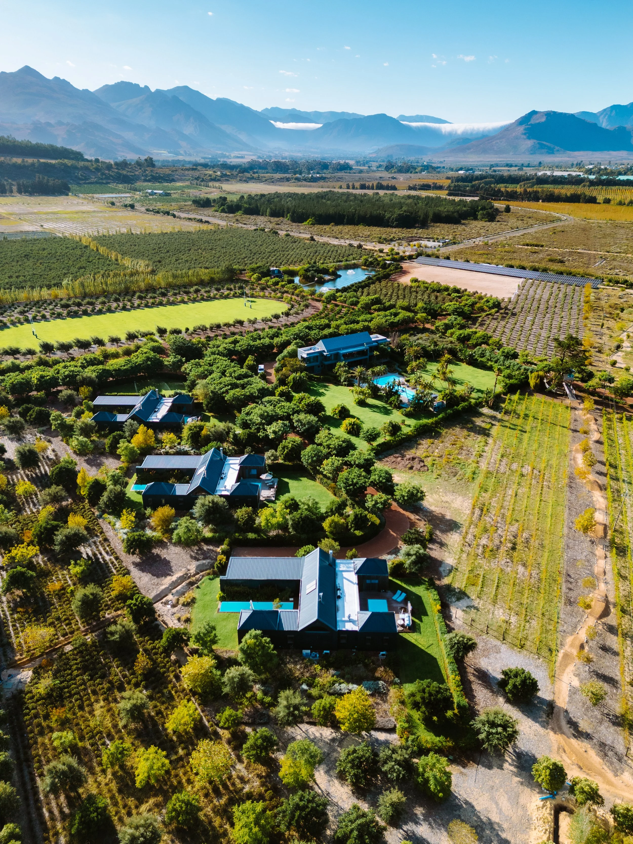 Aerial view of a countryside landscape with multiple modern houses surrounded by trees, pools, and gardens, with agricultural fields and mountains in the background.