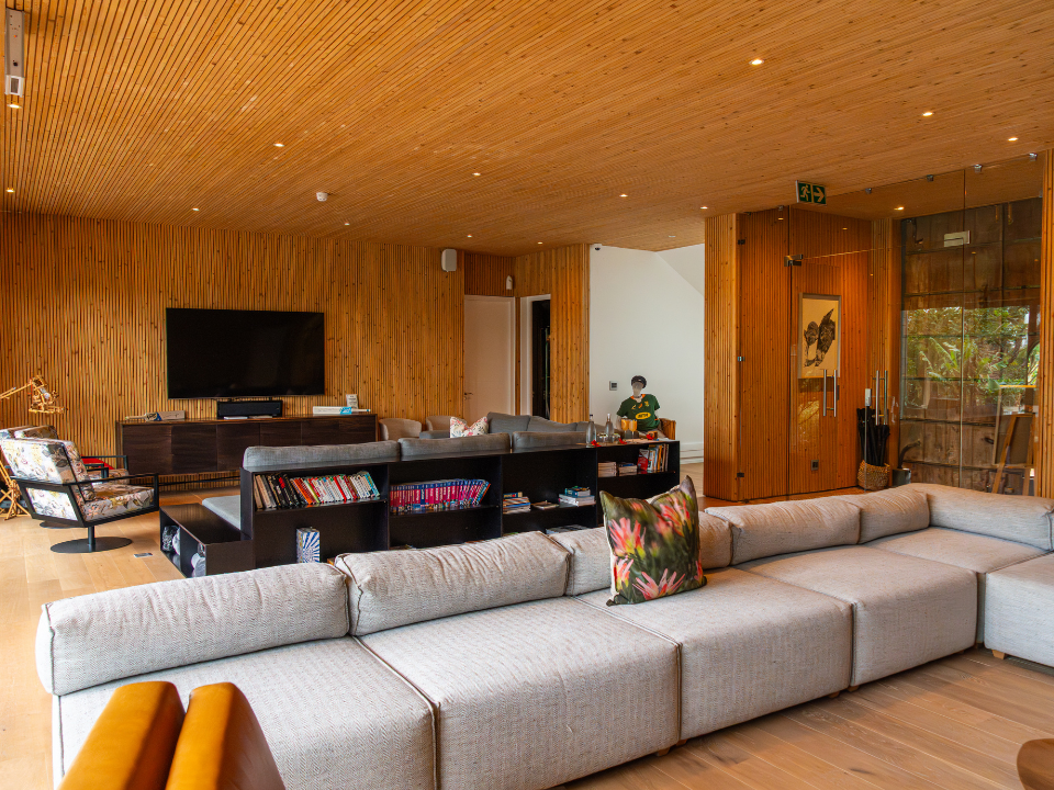 Modern living room with large beige sectional sofa, wooden paneled walls, a flat-screen TV, and a mix of contemporary and floral furniture.