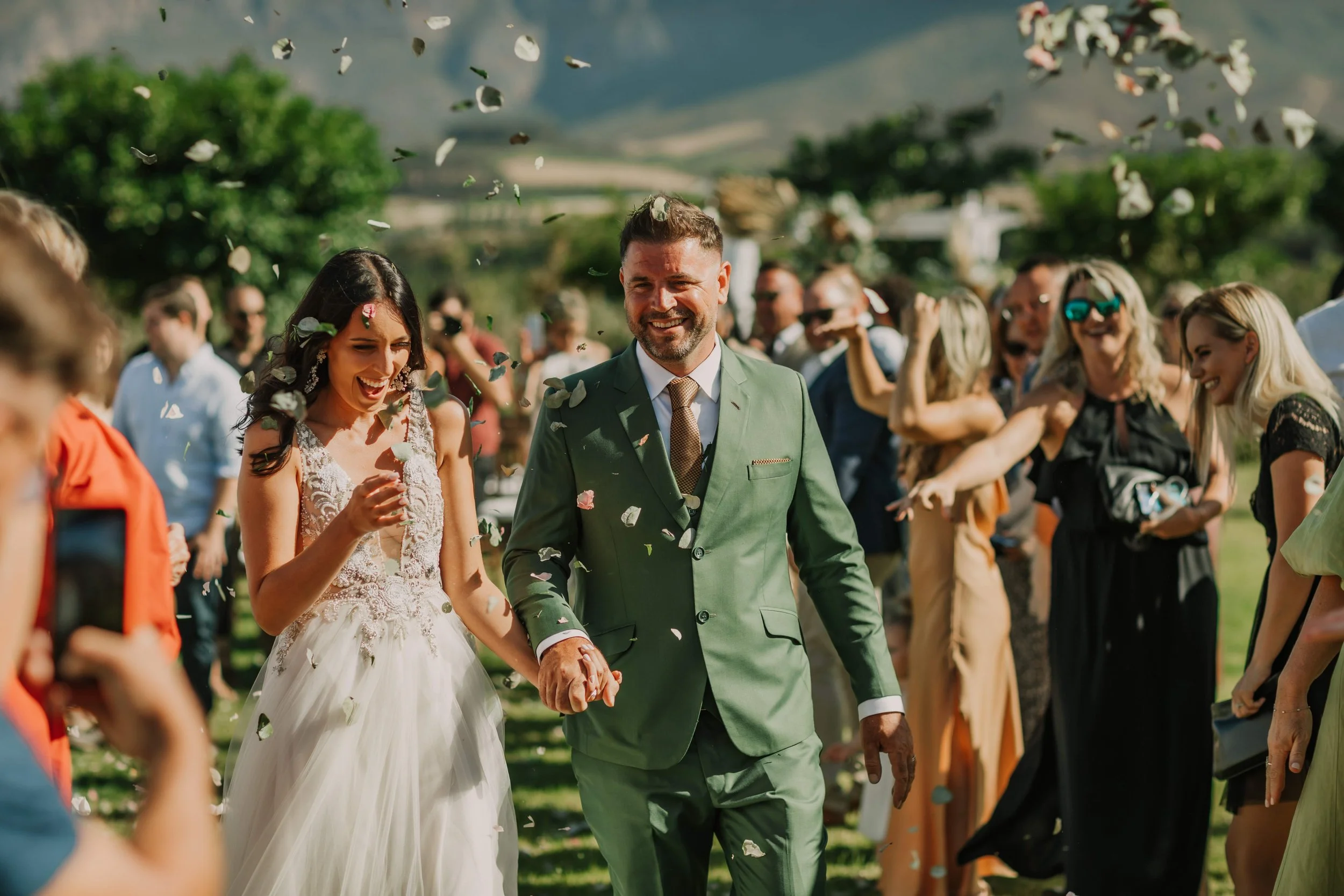 A bride and groom walking hand in hand through a crowd of wedding guests, throwing flower petals during an outdoor wedding celebration on a sunny day.