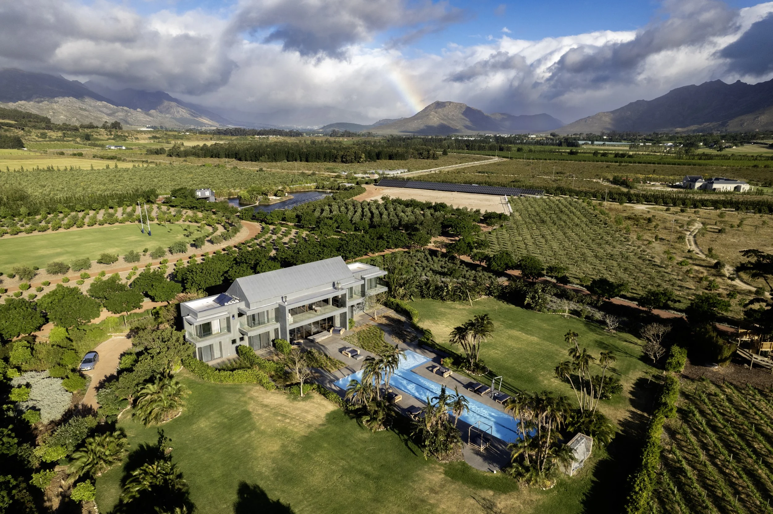 Aerial view of a large modern house with a swimming pool, surrounded by lush green gardens and trees, with fields, vineyards, a river, mountains, and a rainbow in the background.