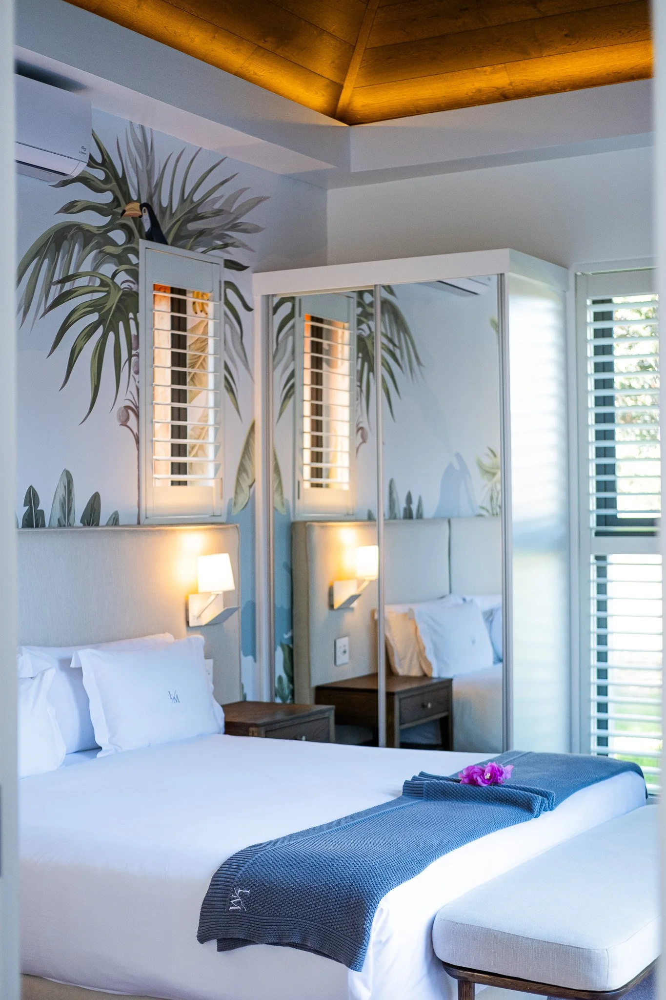 A hotel room with a large bed, white sheets, pillows, a dark gray blanket with a monogram, a flower on the bed, and bedside tables with lamps. The wall above the bed has tropical plant and toucan mural art, and a mirrored closet with sliding doors re