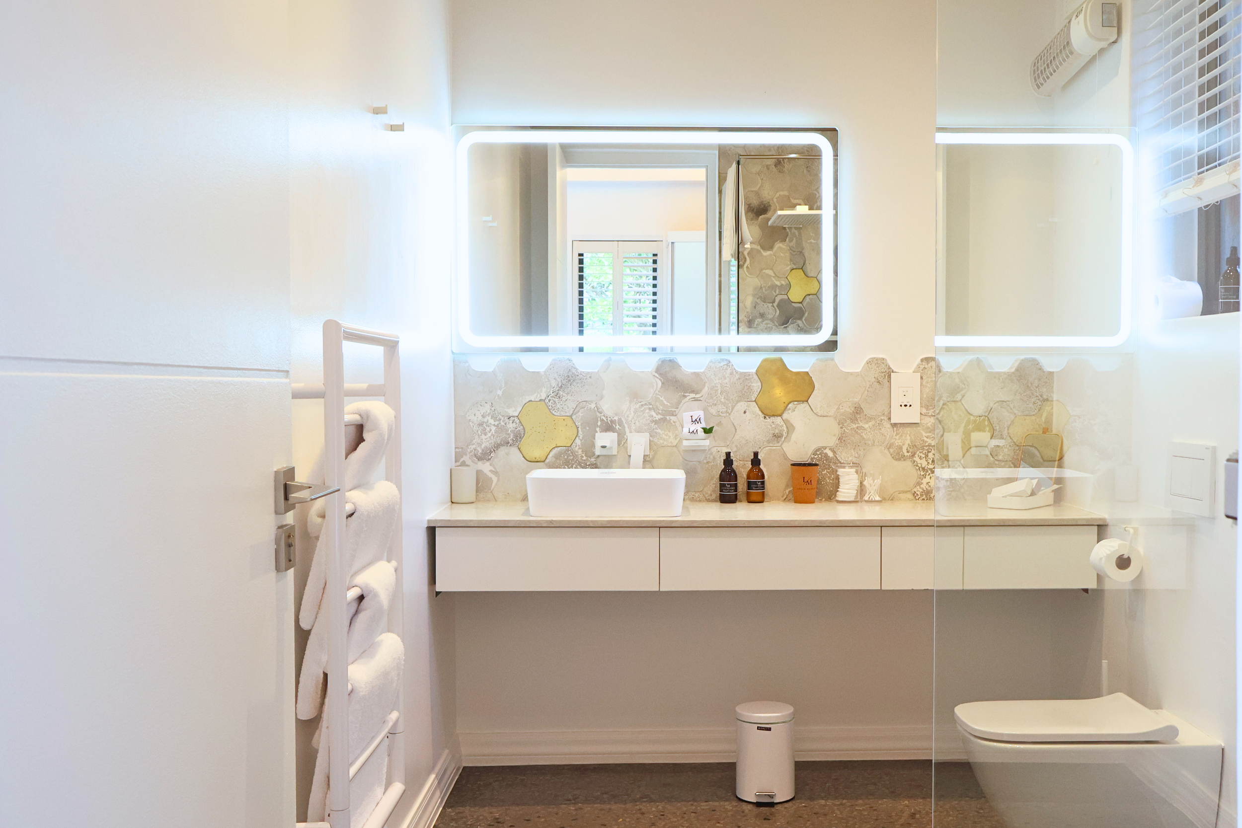 Modern bathroom with a rectangular illuminated mirror, vanity with toiletries, window with shutters, and a white toilet.