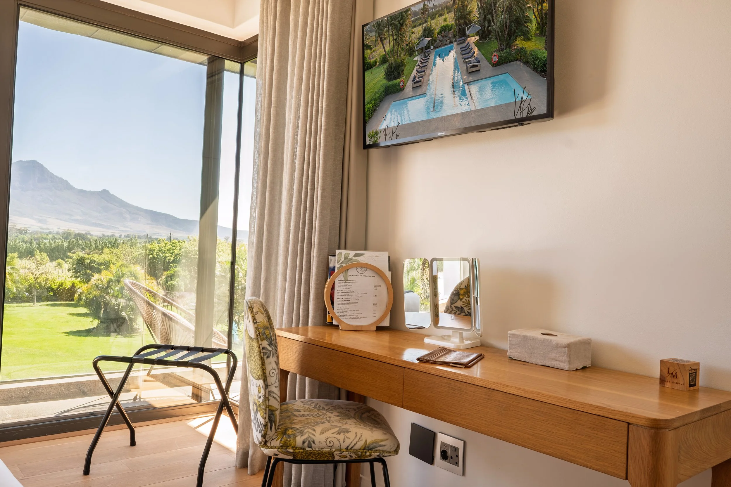 A room with a large window overlooking greenery and mountains. A wooden desk with mirrors, a small desk lamp. A chair with floral upholstery and another chair with a patterned cushion at Ludus Magnus 5 star Luxury Boutique Hotel and Villas