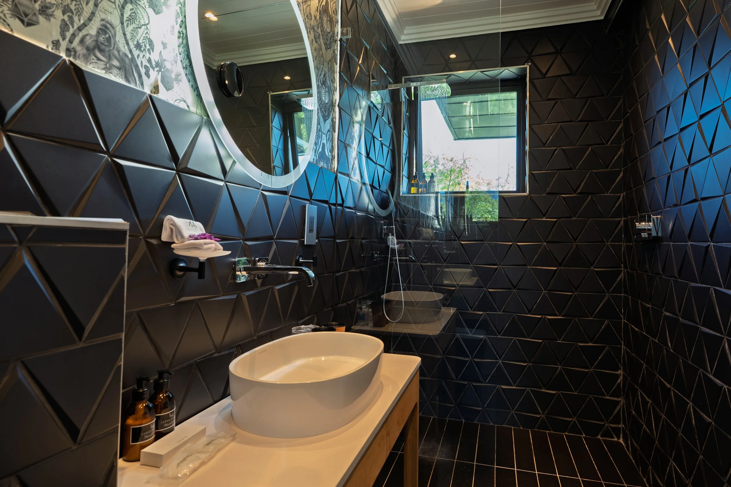Modern bathroom with black geometric tiled walls, a large round mirror, a wooden vanity with a vessel sink, two amber soap dispensers, and a window overlooking greenery.