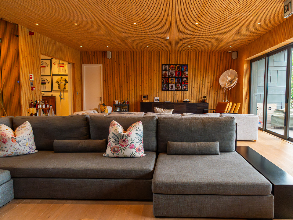 Modern living room with wooden ceiling and wall paneling, beige sectional sofa with floral and solid-colored pillows, sliding glass door to an outdoor patio, and contemporary decor including artwork and a large fan.