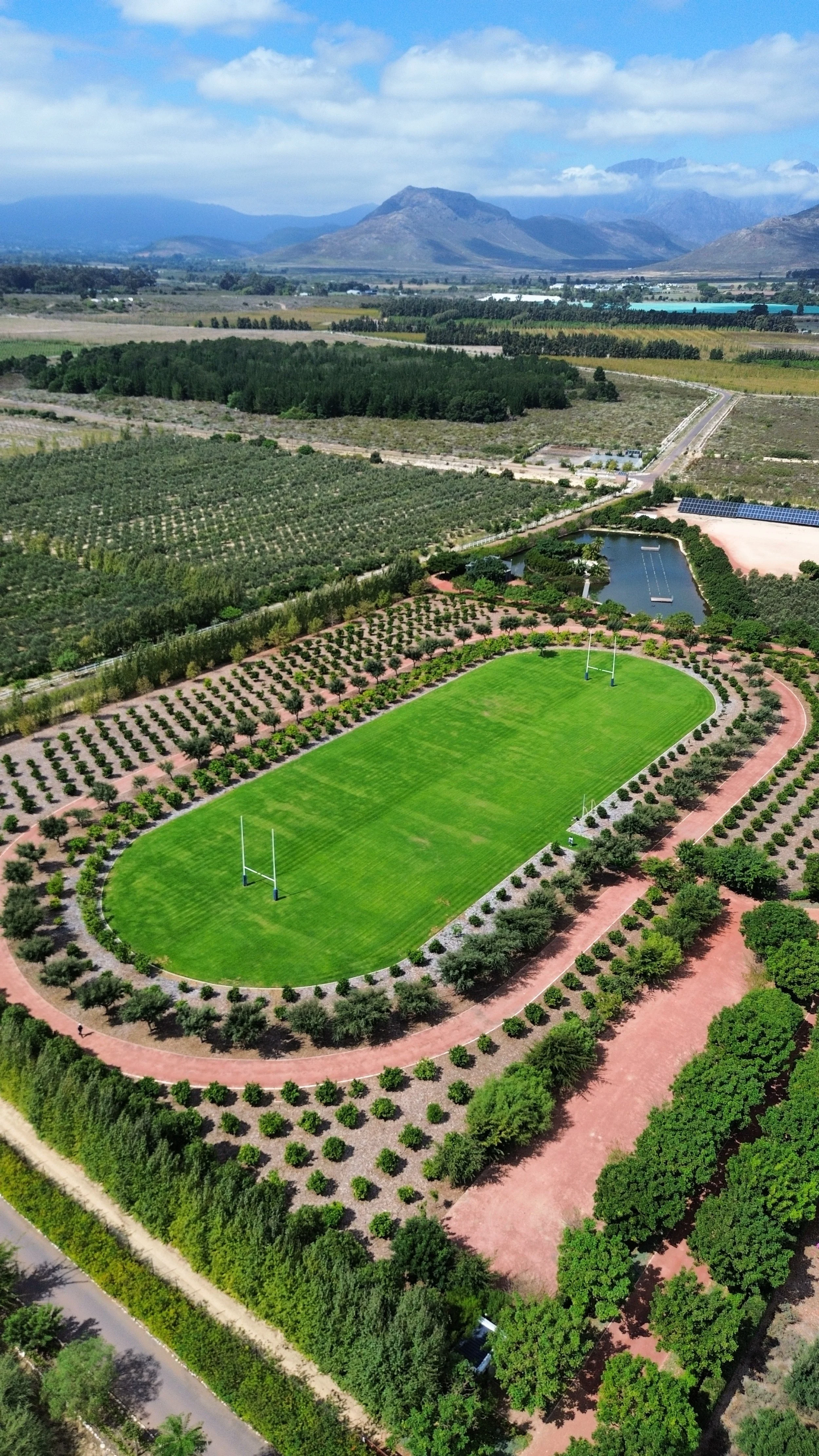 An aerial view of a lush green rugby field surrounded by trees and pathways, with mountains and farmland in the background.
