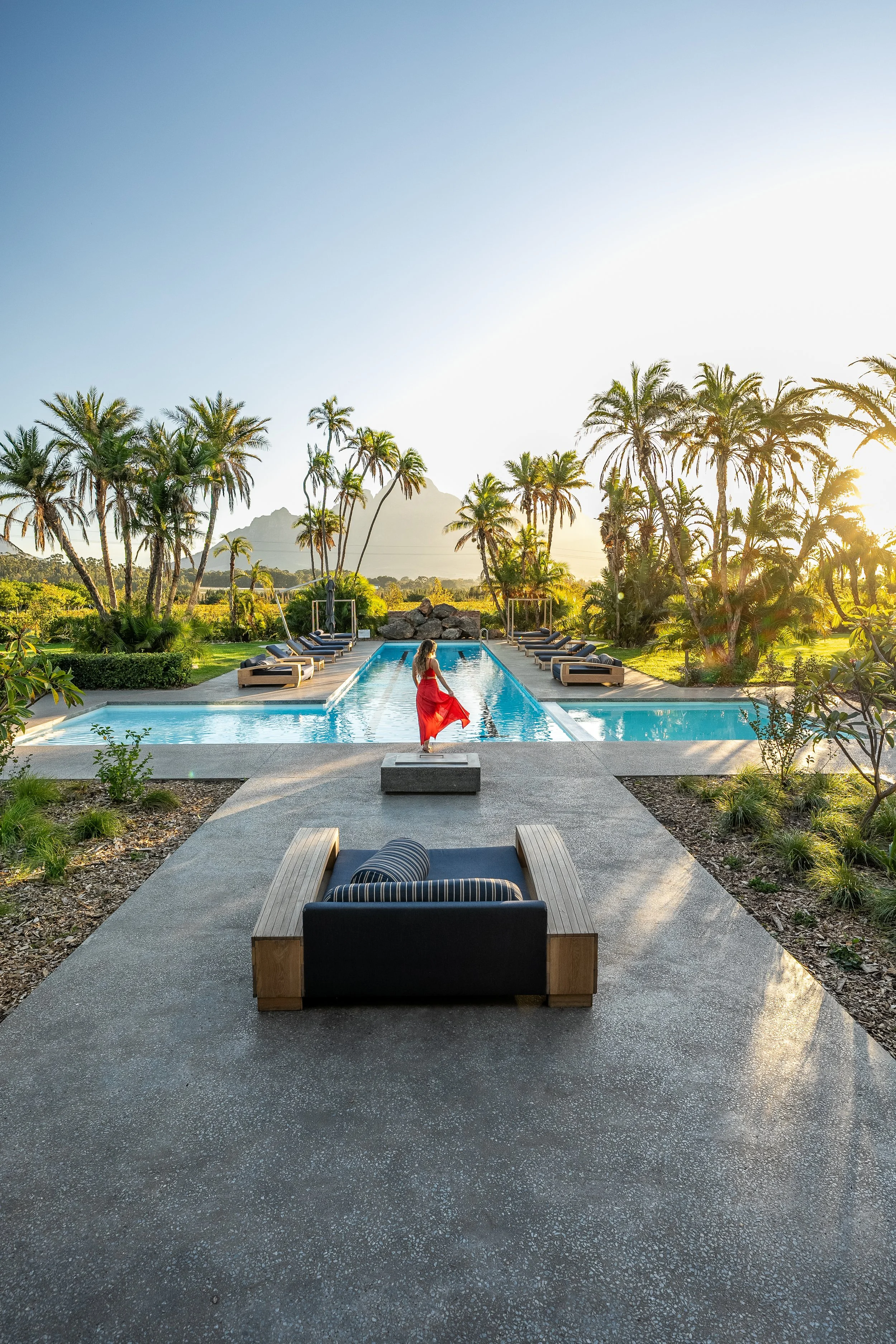 A woman in a red dress walking along the edge of a luxurious swimming pool surrounded by palm trees at sunset, with lounge chairs along the poolside and mountains in the background.