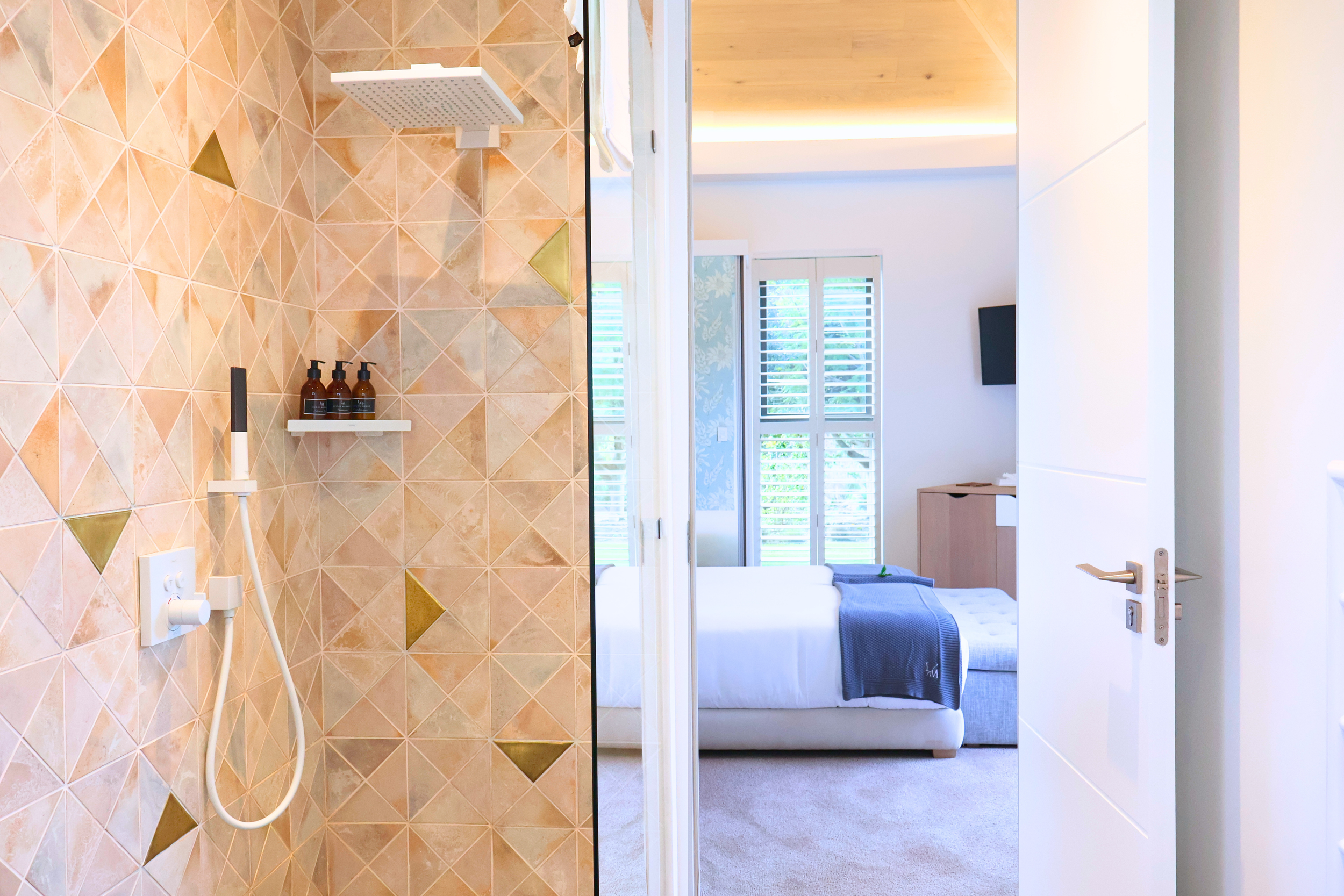 A bathroom shower area with beige and gold patterned tiles, a white showerhead, and three soap or shampoo bottles on a shelf. The shower area is open to a bedroom, which has a white bed with a dark throw blanket, large windows with blinds, and light-