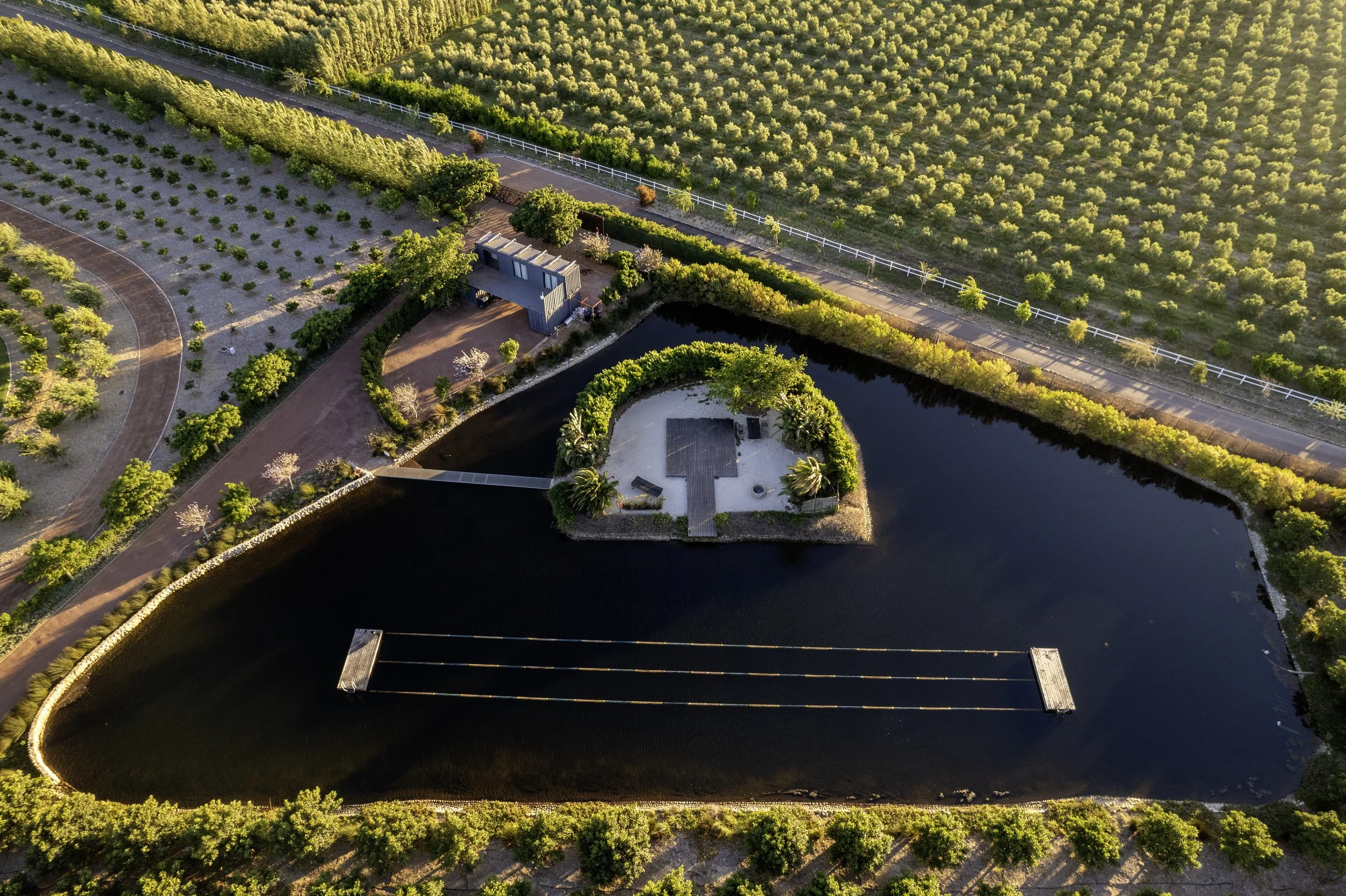 Aerial view of a park with a small dock on a pond, surrounded by trees, a walking path, and a large orchard in the background.