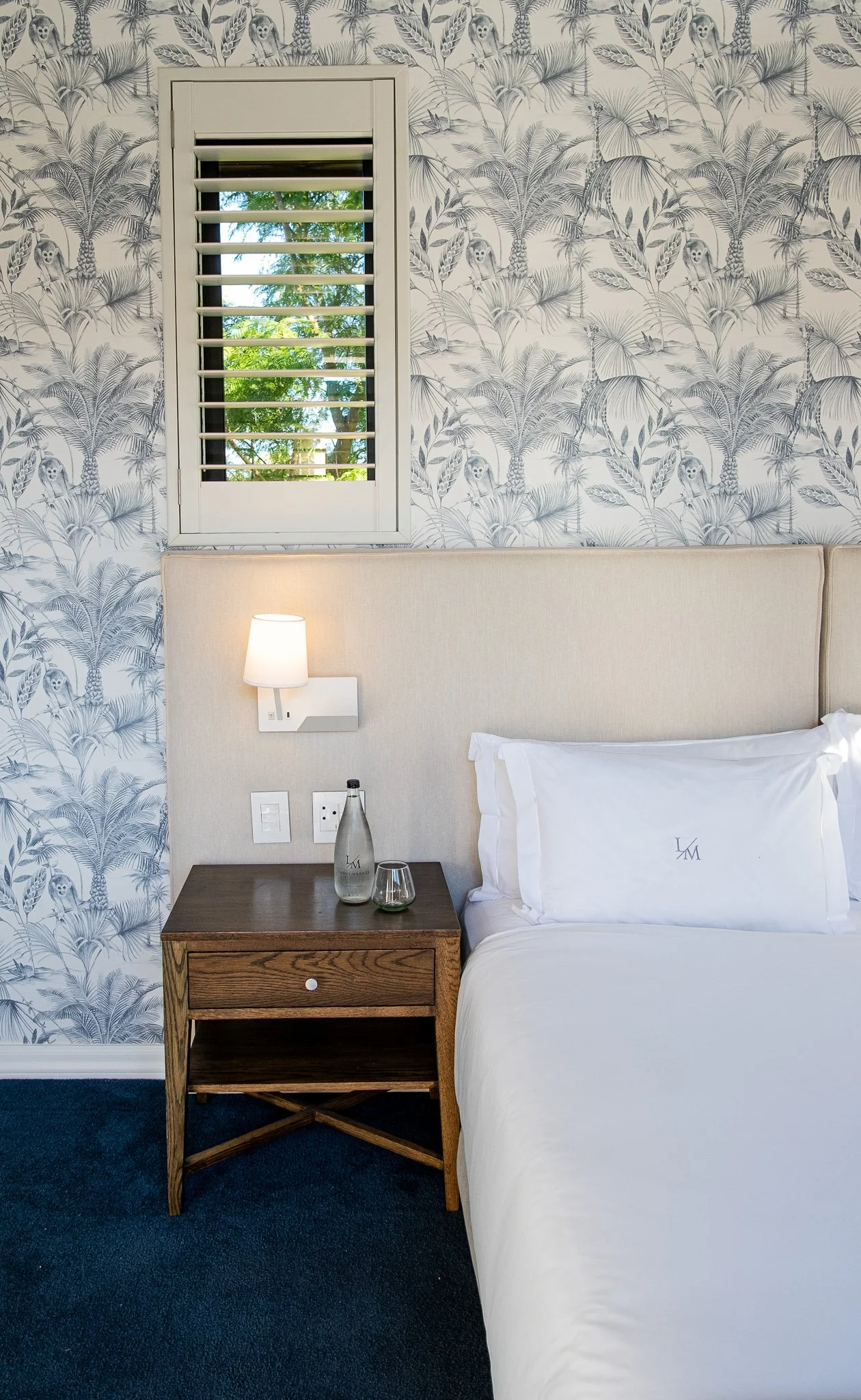 Hotel room with a bed, wooden nightstand, white pillow with embroidered initials, wall-mounted light, window with white shutters showing greenery outside, tropical wallpaper with animal illustrations, and a bottle of water on the nightstand.