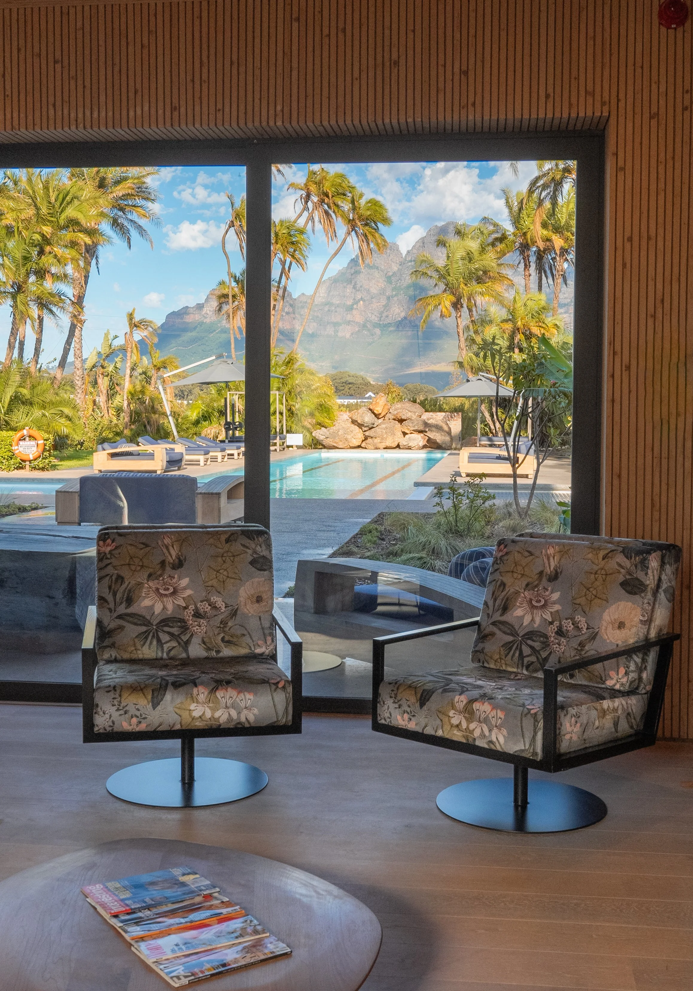 Indoor view of a lounge area with two floral upholstered chairs facing large glass doors. Outside, an emerald-green swimming pool is surrounded by lounge chairs, umbrellas, palm trees, and a mountain backdrop under a partly cloudy blue sky.