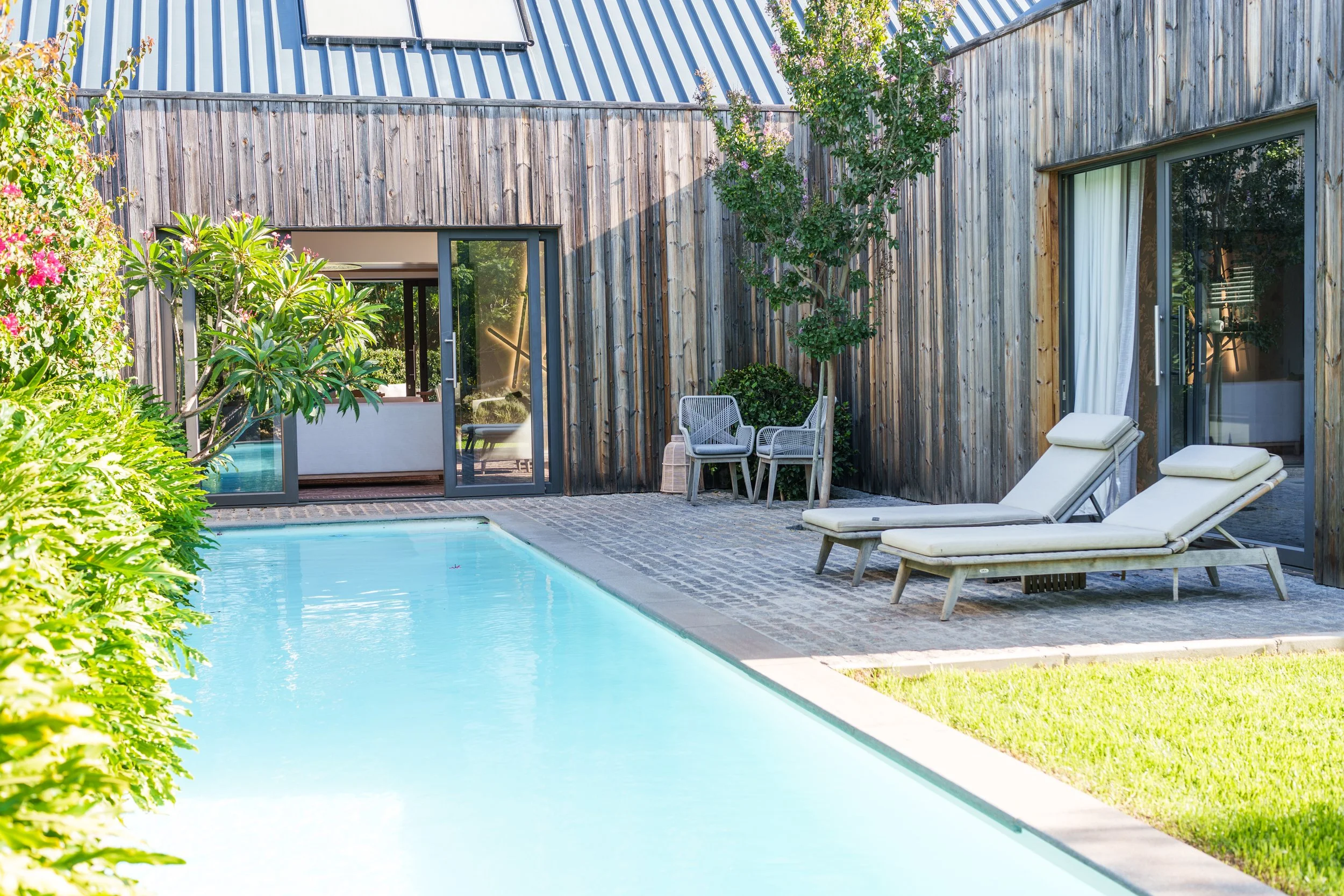 Backyard with a swimming pool, wooden house walls, patio furniture, and surrounding greenery.