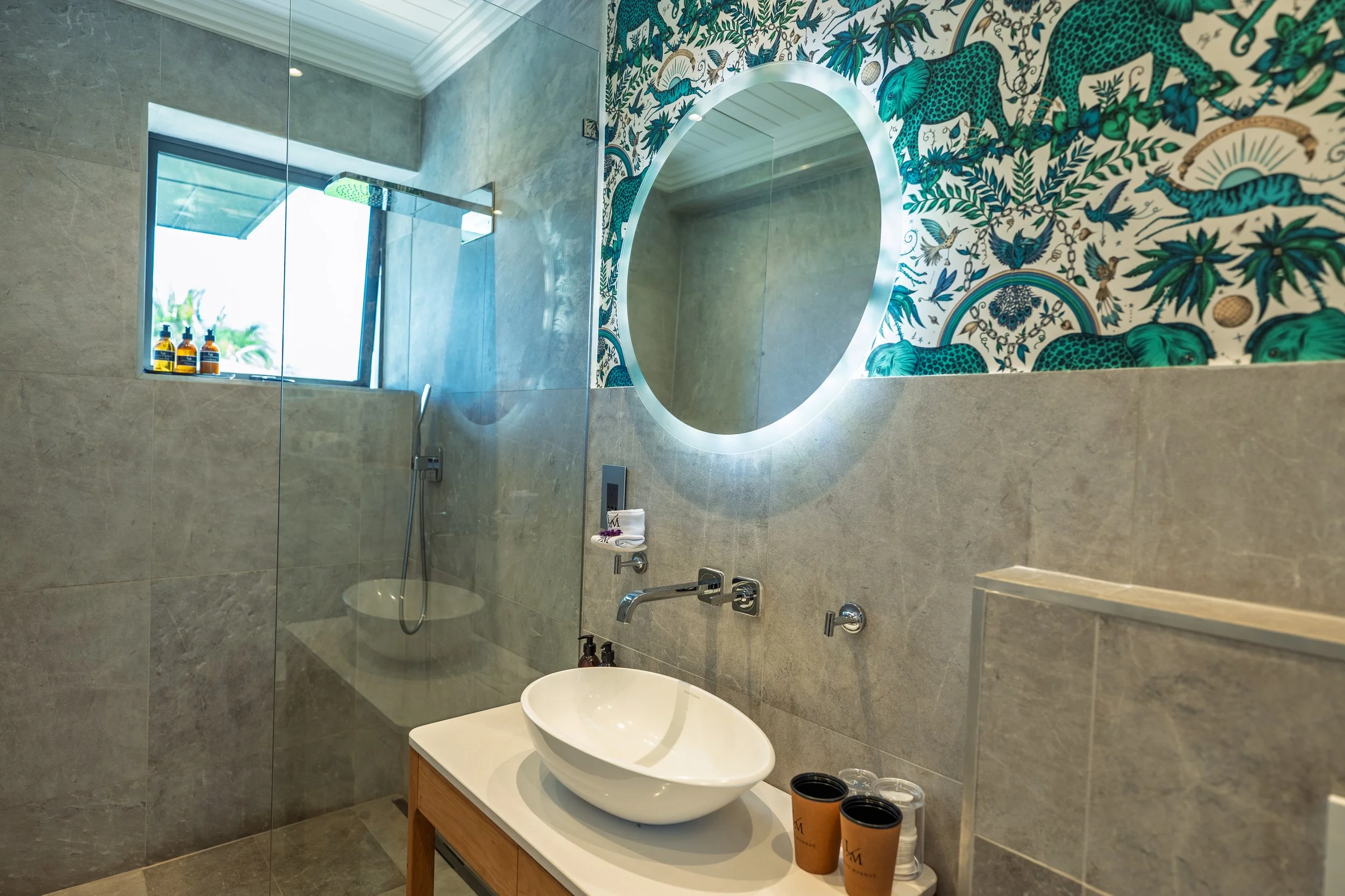 Modern bathroom with a walk-in shower that has a window and bottles of toiletries on the sill, a round illuminated mirror, a vessel sink on a wooden vanity with cups, and a decorative feature wall with jungle-themed artwork of leopards, snakes, and b