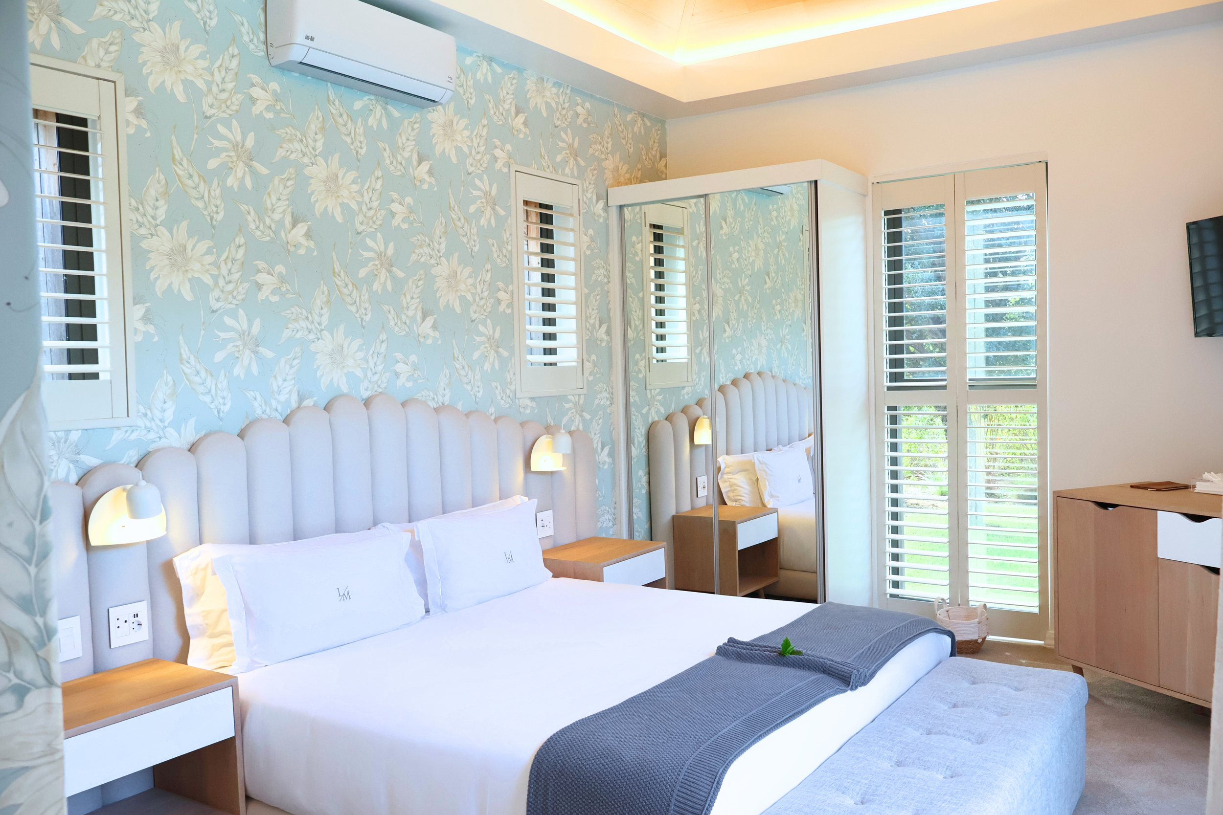 A modern bedroom with a floral blue wallpaper, white bed with pillows, bedside tables, wall-mounted lamps, large window with white shutters, a mirror, a television, and an air conditioning unit.