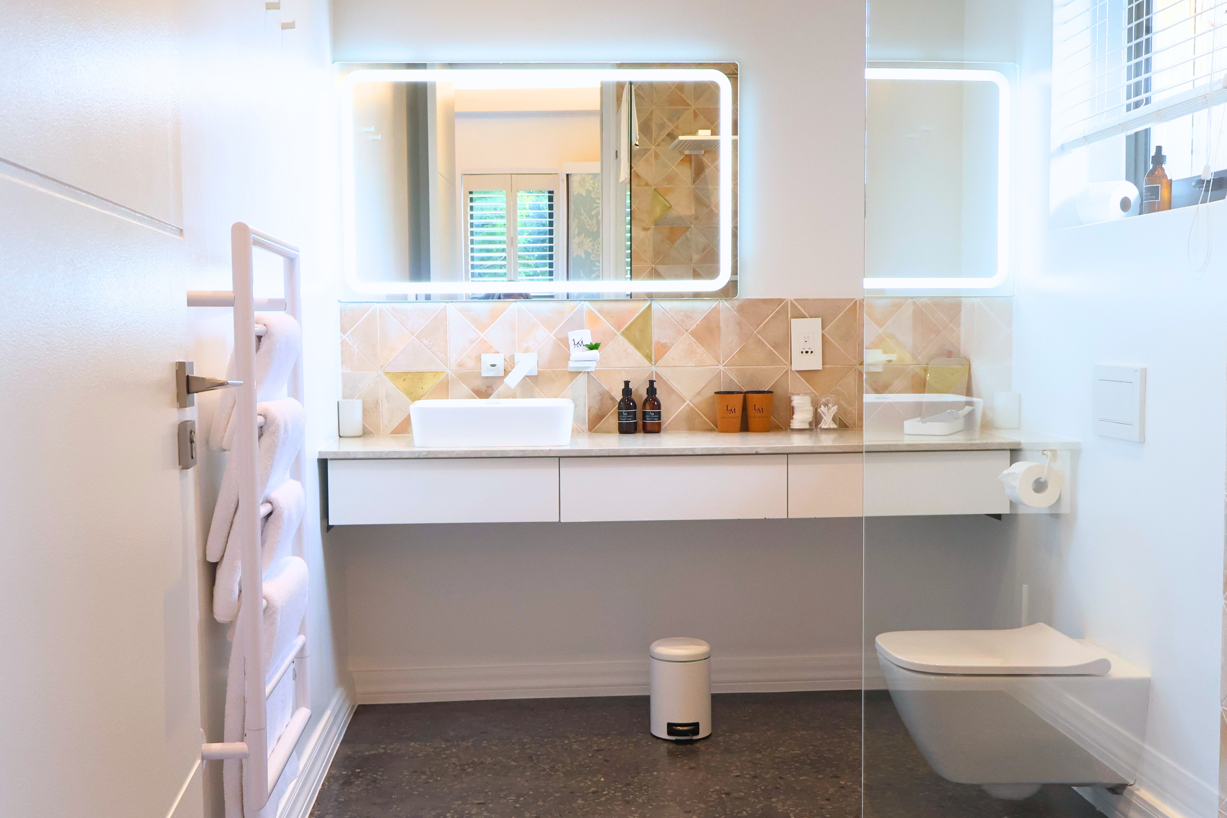 Clean and modern bathroom with a wall-mounted mirror with LED lights, a white sink with accessories, a toilet, and a window with blinds, with a small trash bin on dark speckled floor.