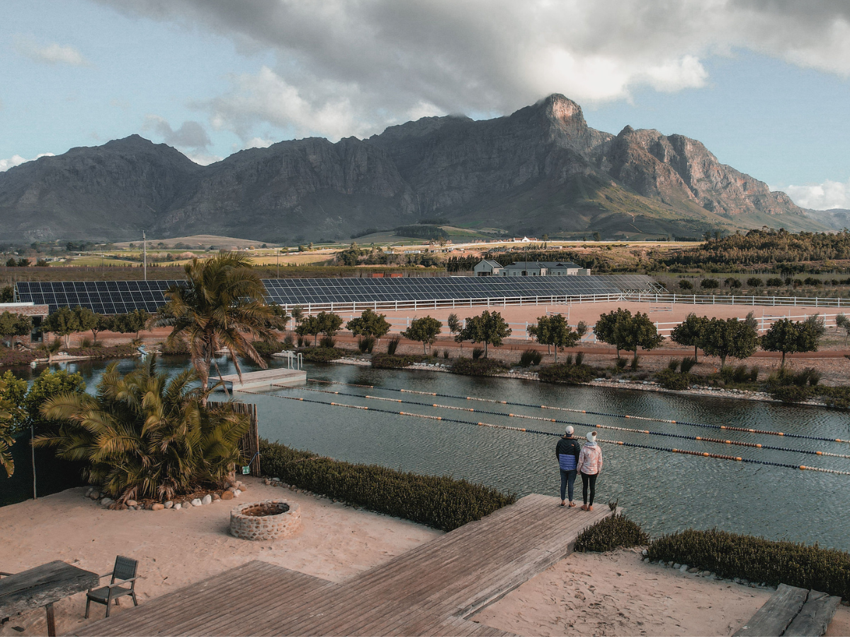 Two people standing on a wooden dock looking at a pond or lake with solar panels and trees, mountains in the background, cloudy sky.