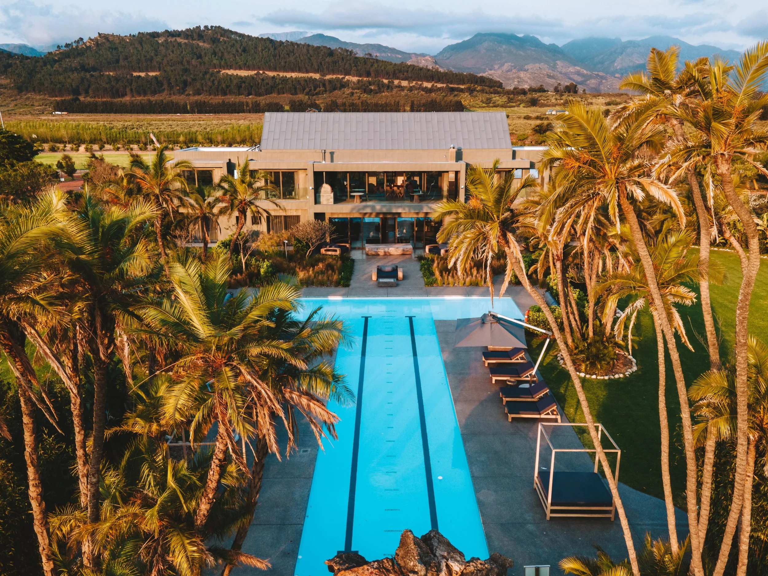 Aerial view of a luxury house with a swimming pool, surrounded by palm trees and mountains in the background.