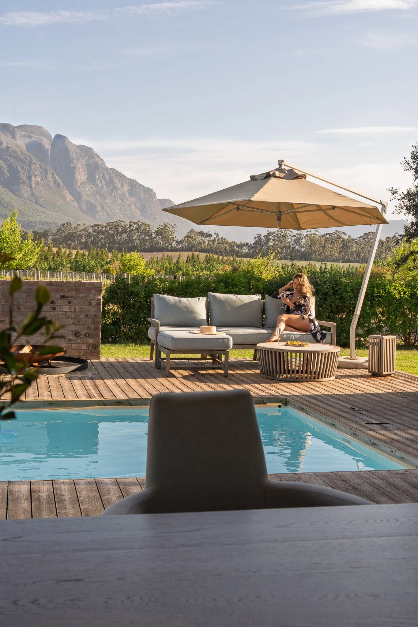 A woman relaxing on a patio sofa under a large beige umbrella, next to a swimming pool, with mountains and greenery in the background, during sunset.