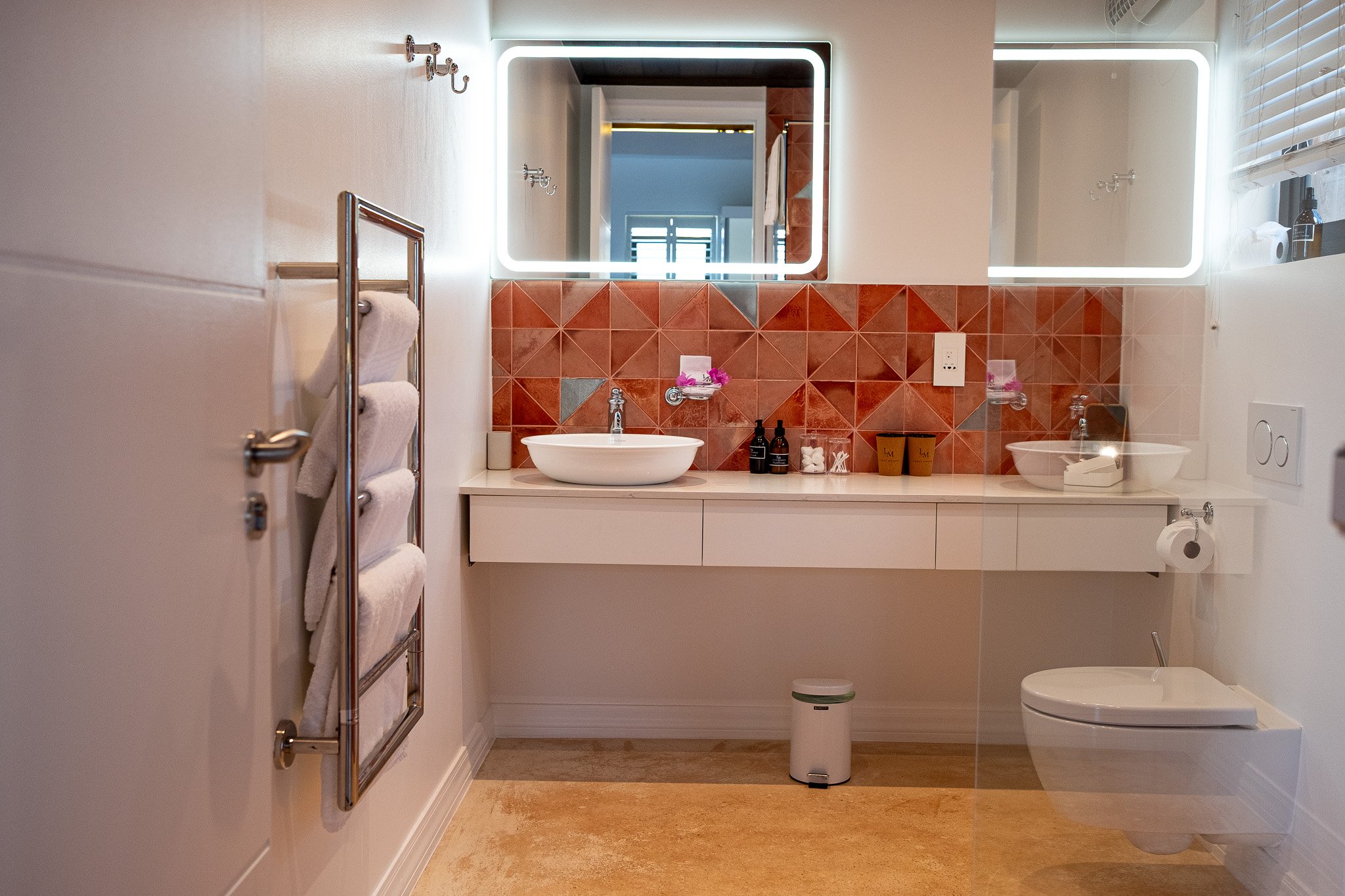 A modern bathroom with a white counter, two vessel sinks, and illuminated mirror. There are gray and brown tiles on the wall behind the sinks. To the left, there is a towel rack with white towels and hooks on the wall above. On the right, there is a 