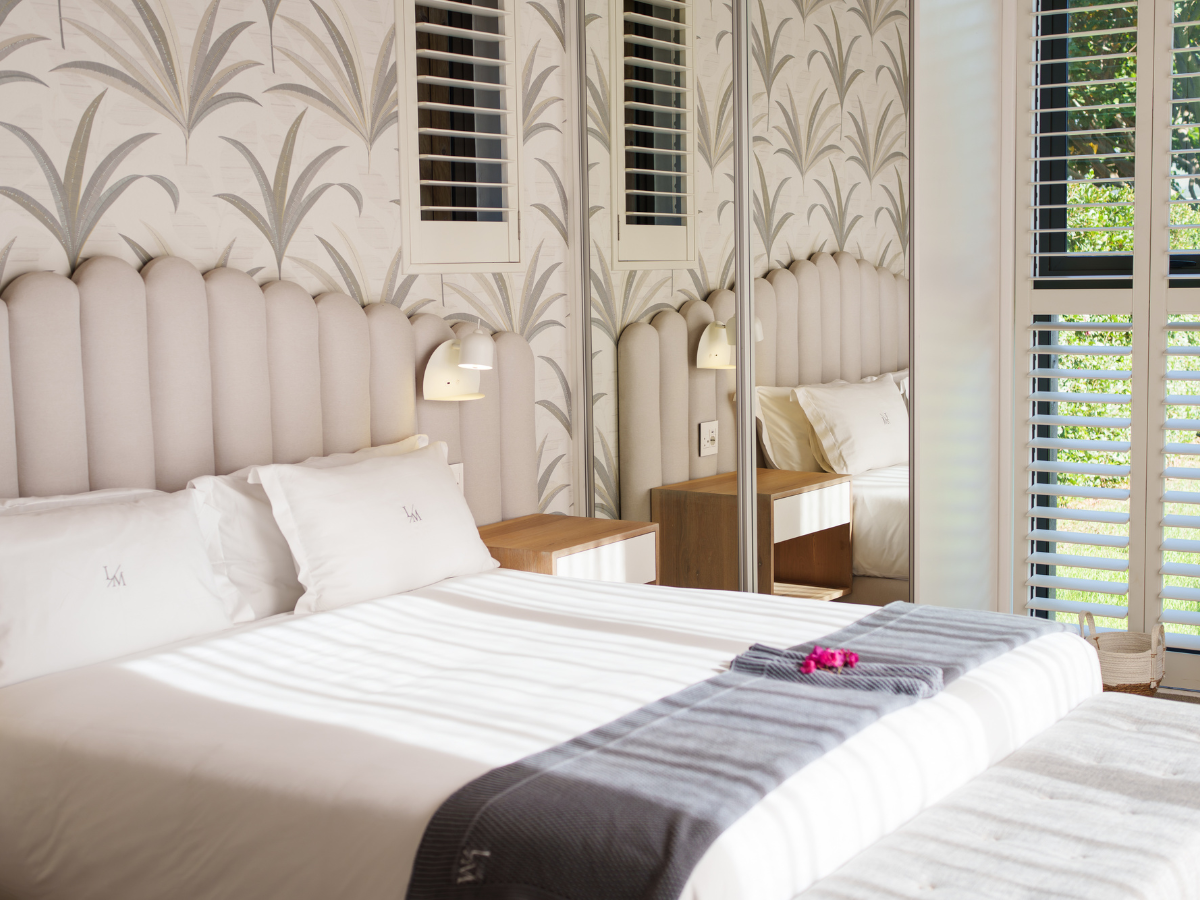 A neatly made bed with white linens and pillows, flanked by two wooden bedside tables with wall-mounted lamps, mirror closet doors, patterned wallpaper, and a large window with blinds letting in natural light.