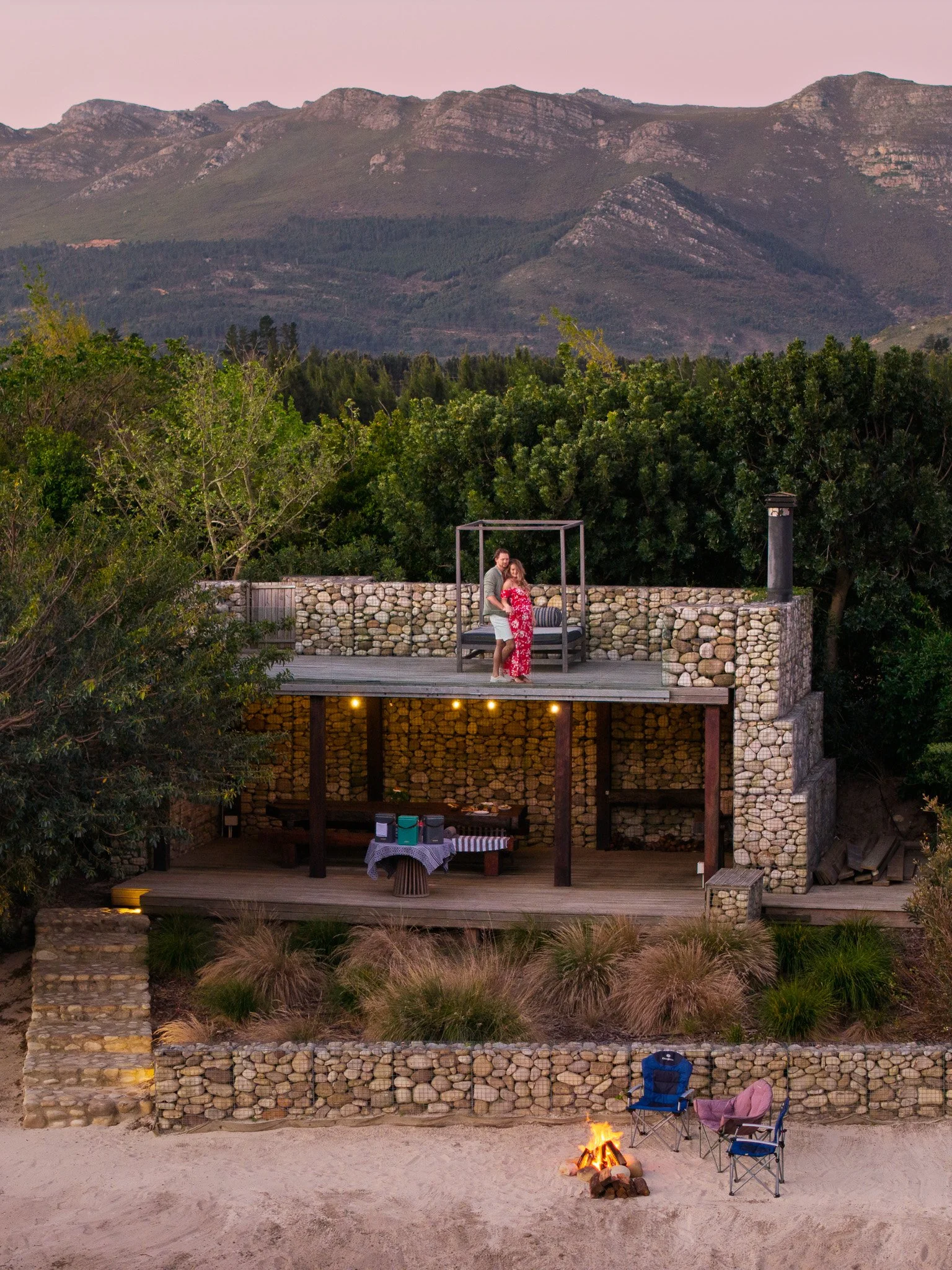 A couple standing on an outdoor deck with a canopy, overlooking a scenic landscape of hills and trees, with a beach and campfire in the foreground.