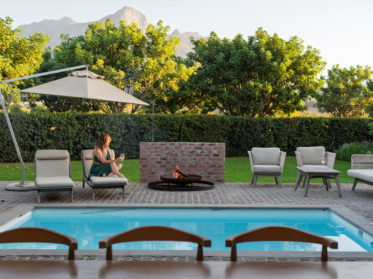 Woman sitting on a lounge chair by a swimming pool, holding a glass of wine, with a brick fire pit, outdoor furniture, trees, and mountains in the background.