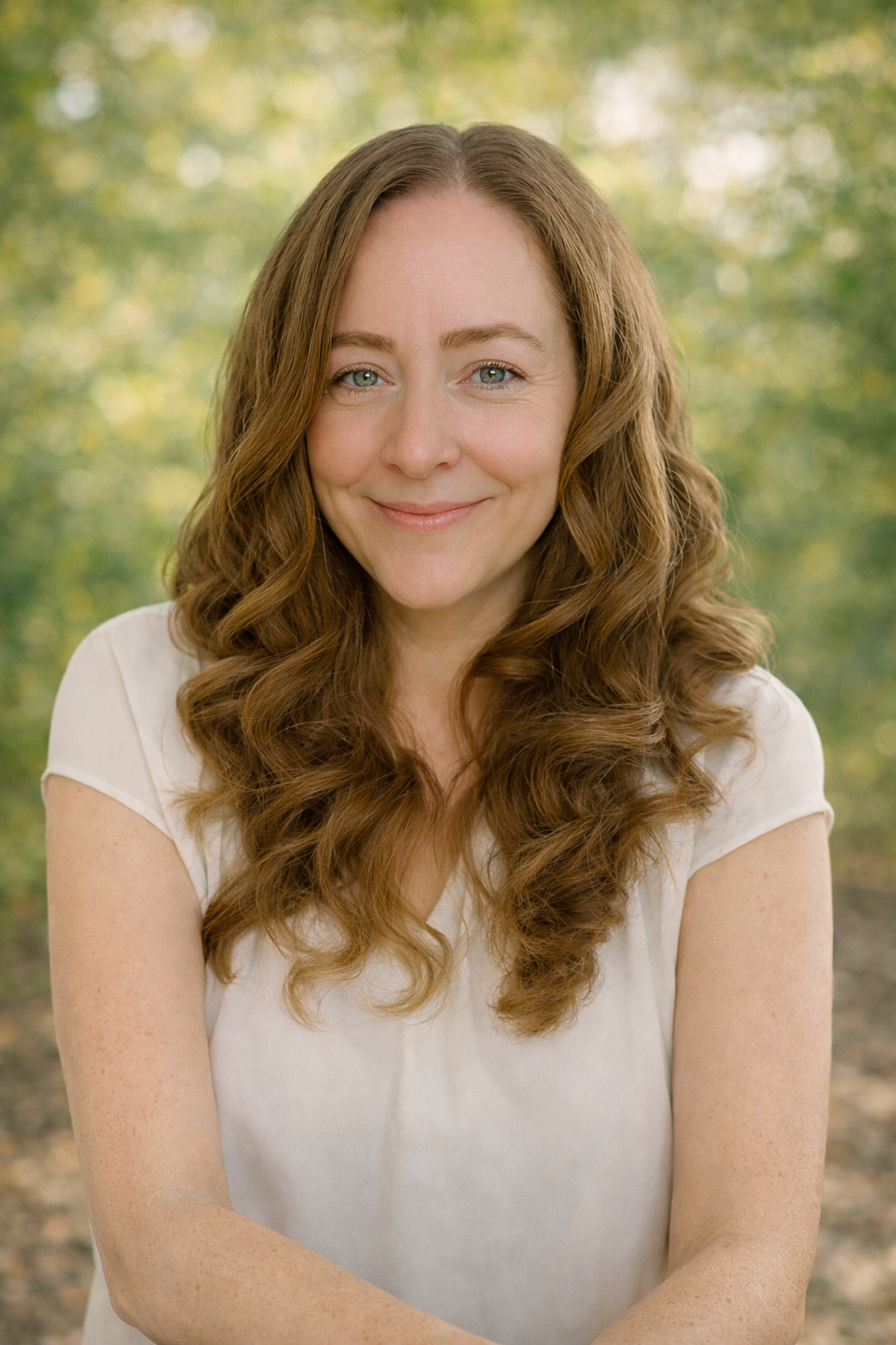 A woman with long, curly, light brown hair smiling outdoors on a sunny day, with blurred green trees in the background.