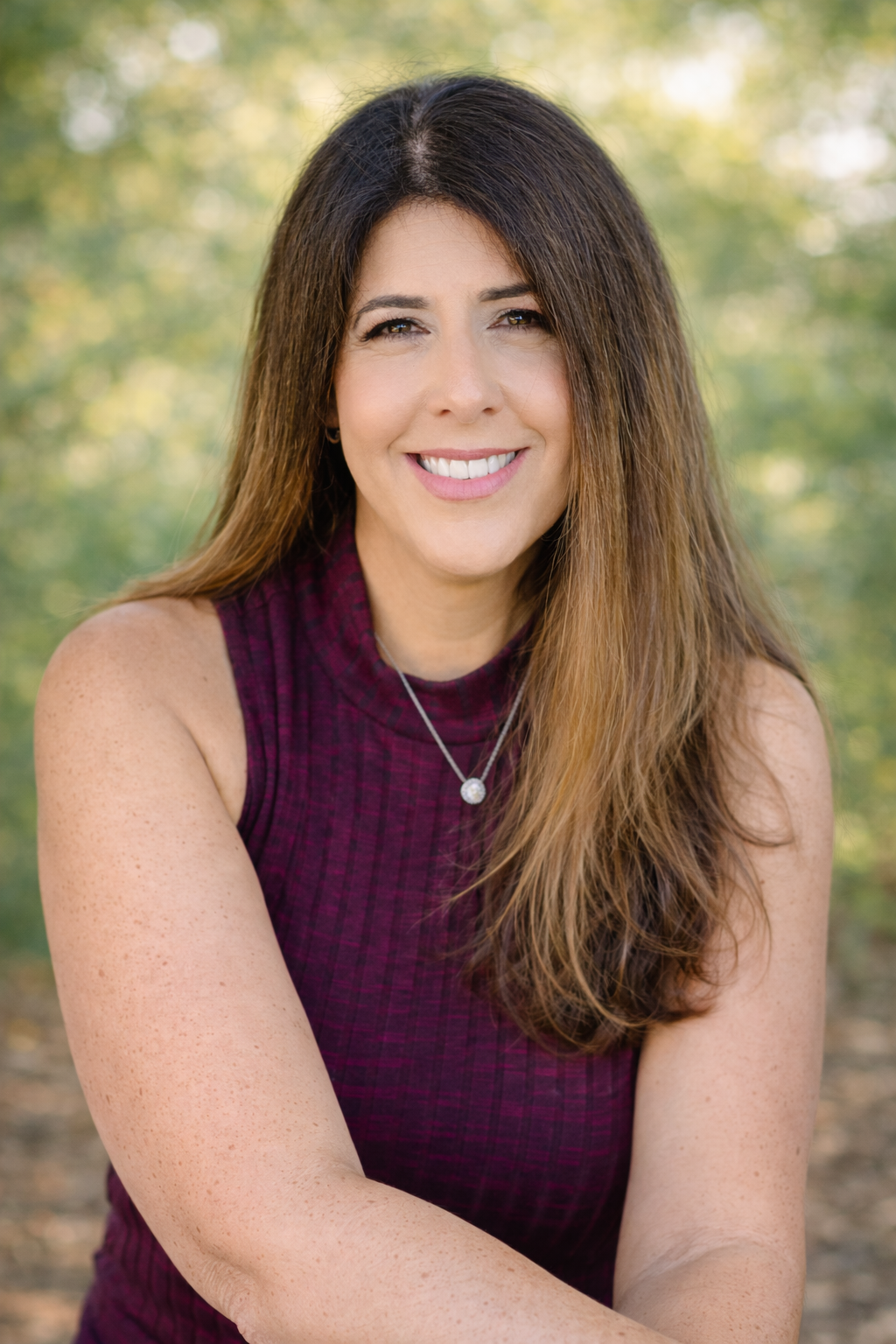 A woman with long brown hair and light skin smiling outdoors, wearing a sleeveless maroon top and a necklace, with a blurred green and yellow background.