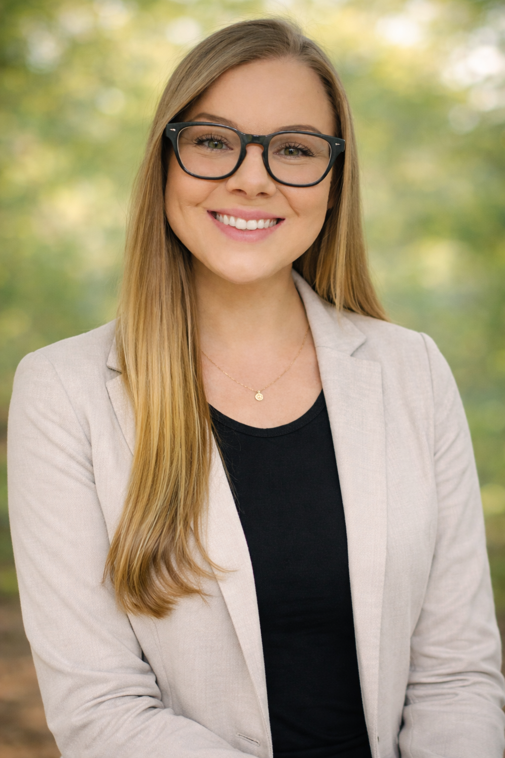 A young woman with long blonde hair, wearing glasses, a beige blazer, and a black top, smiling outdoors with a blurred green, leafy background.