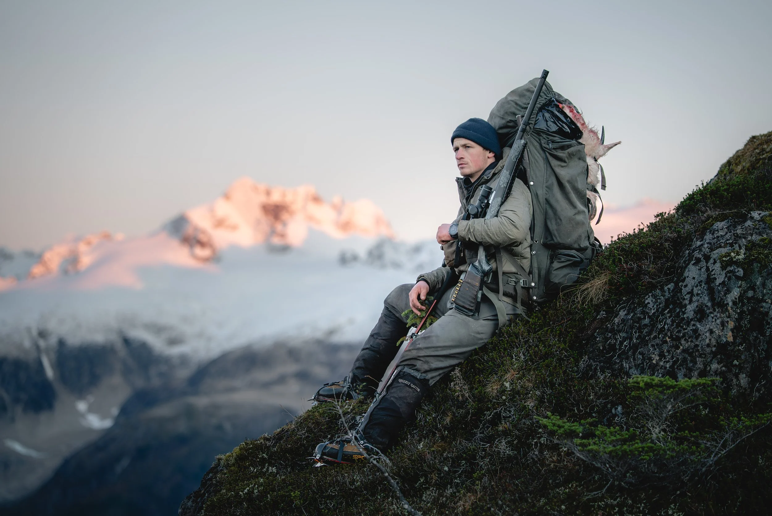 A man sitting on a rocky mountainside wearing outdoor gear, a large backpack, holding a rifle, with snow-capped mountains in the background.