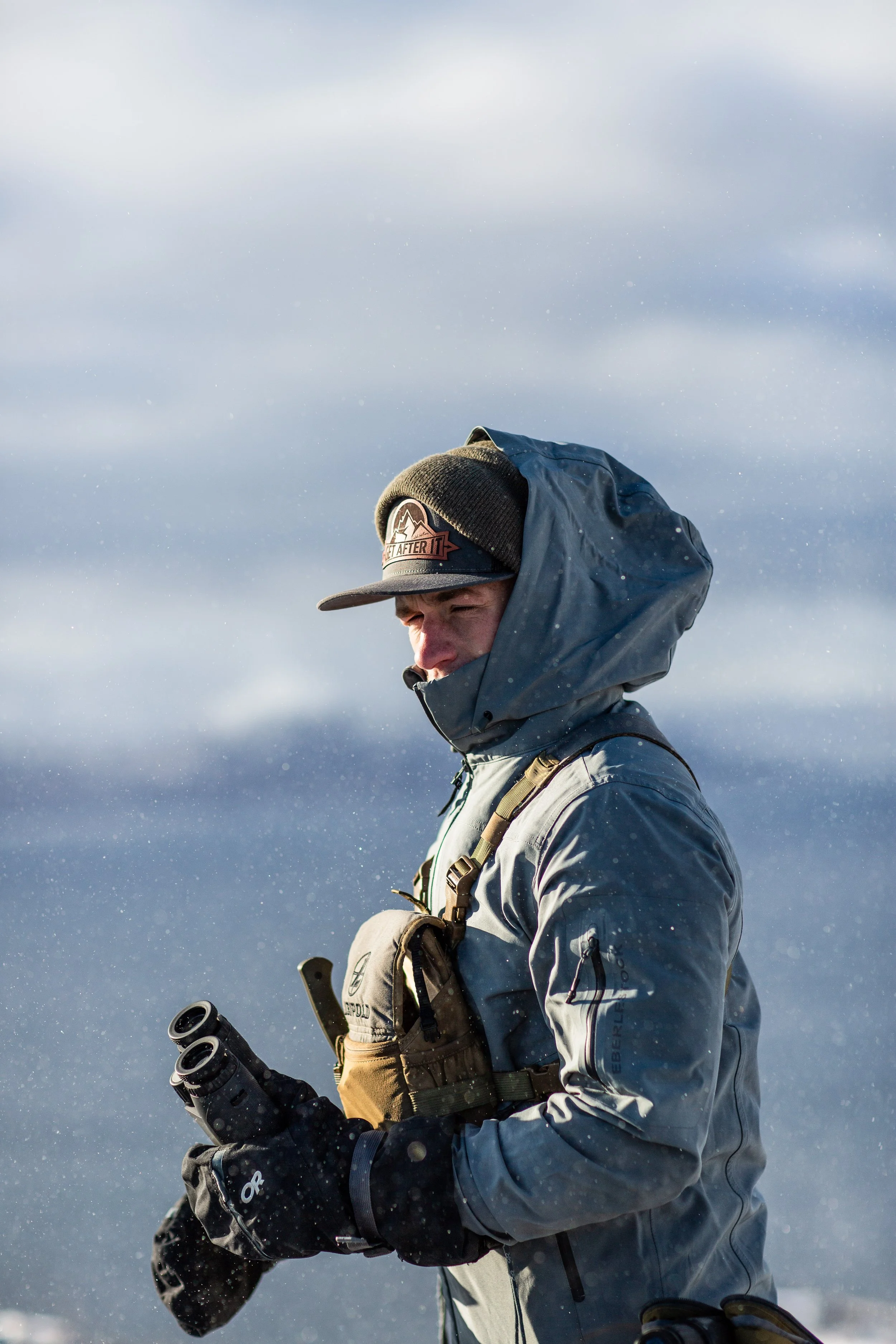 Man dressed in winter outdoor gear with binoculars, standing in a snowy, cold environment.