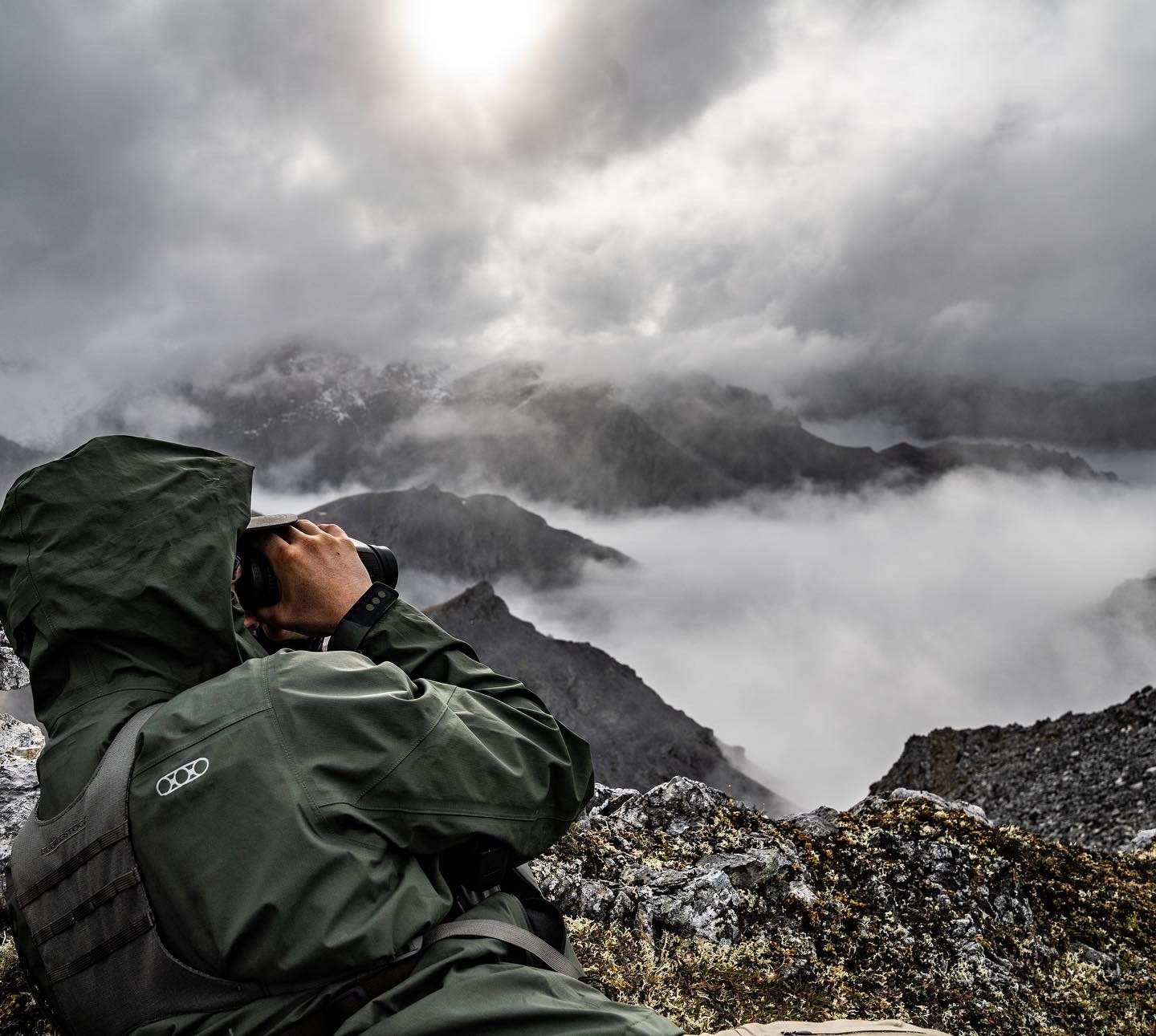 Person wearing waterproof jacket with hood, sitting on rocky terrain, looking through binoculars at a cloudy mountain landscape with mist and rugged peaks.