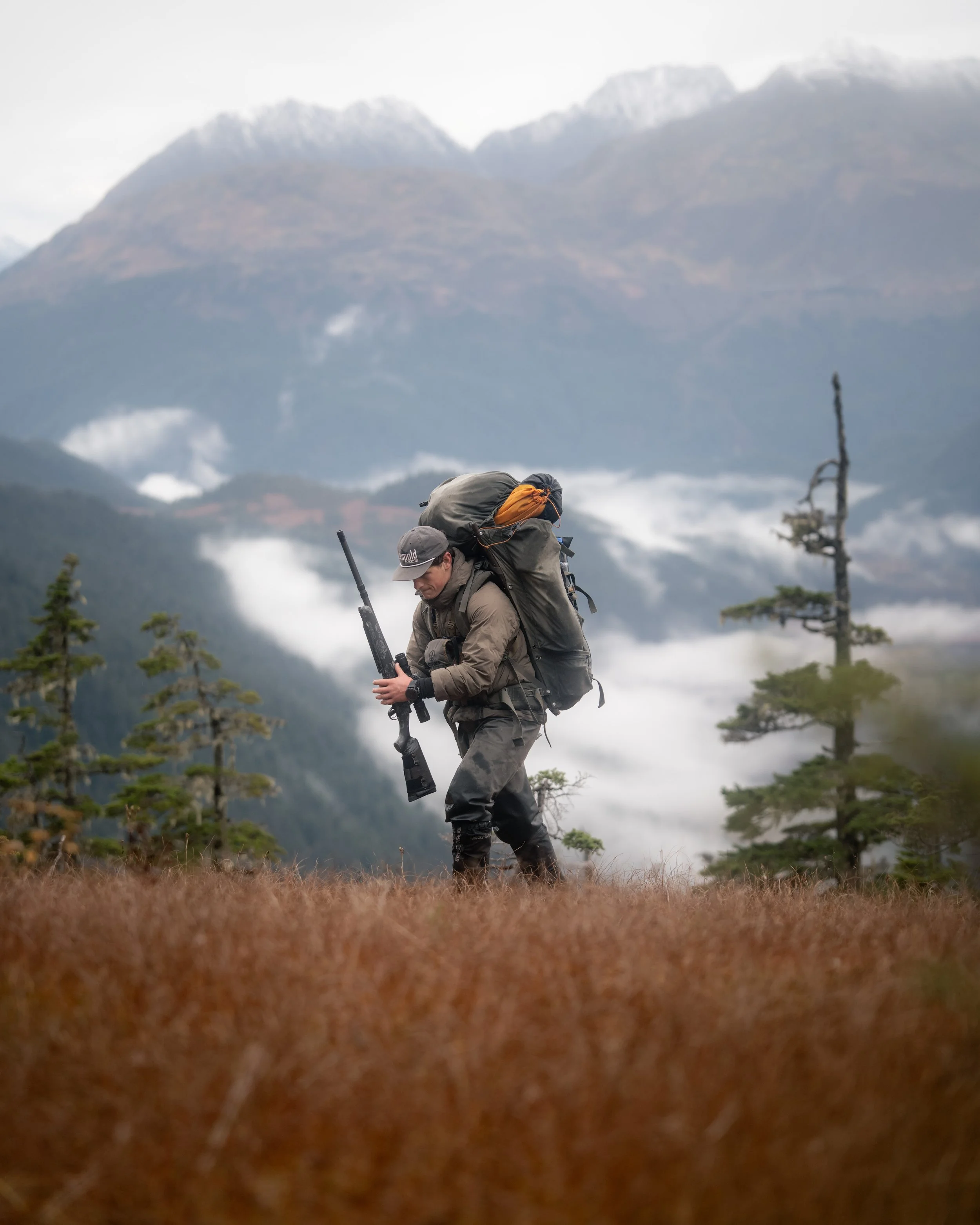 A person dressed in outdoor gear carrying a large backpack and rifle, walking through a grassy field with trees and mountains in the background.