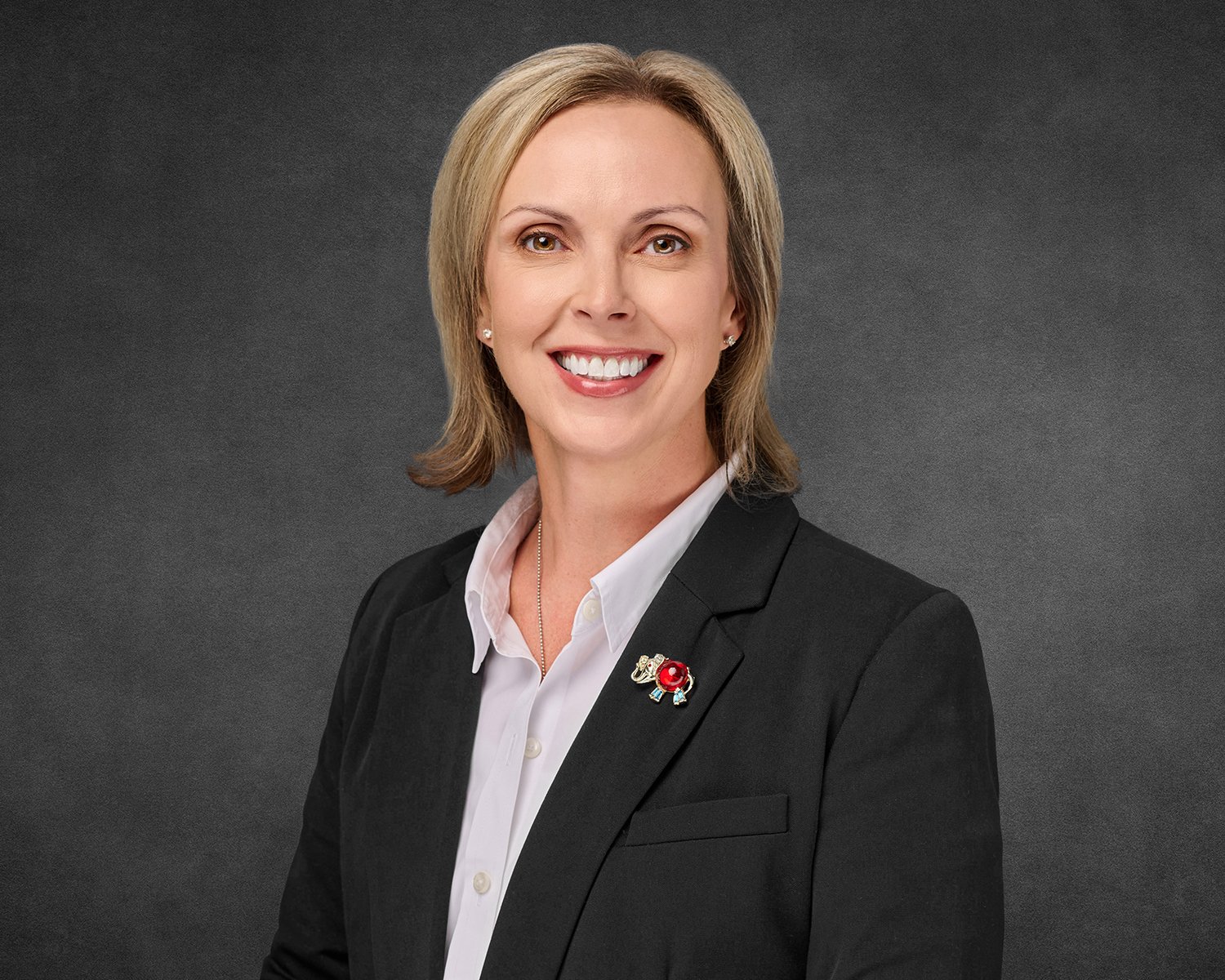 A professional headshot of woman with short blonde hair, wearing a black blazer over a white shirt, smiling at the camera against a dark gray background.