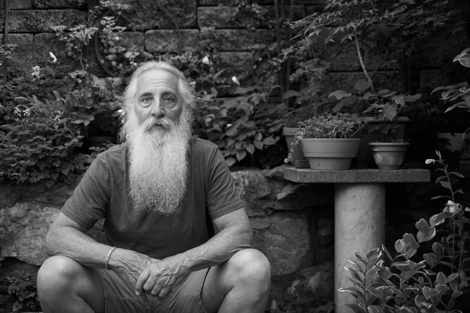 An elderly man with a long white beard and hair sitting in a garden with plants and a stone wall behind him.
