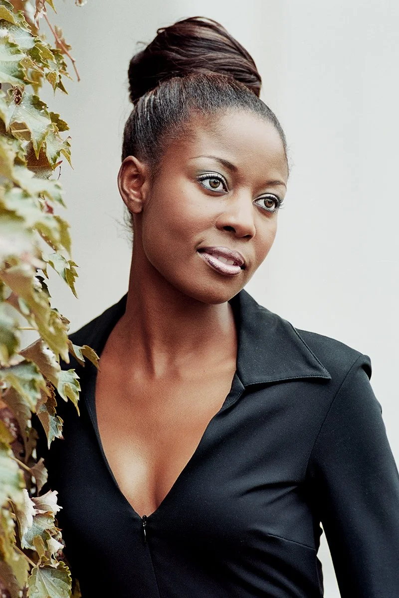 A woman with dark skin and hair styled in an elegant updo, wearing a black top, standing next to green ivy leaves.