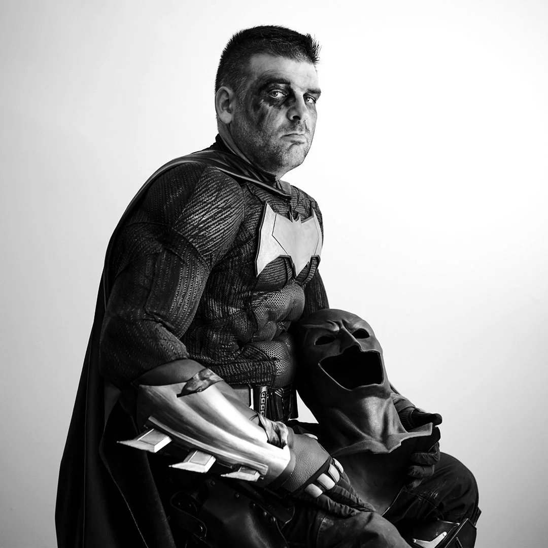 Black and white photo of a man dressed as Batman holding a mask with an exaggerated scream expression.