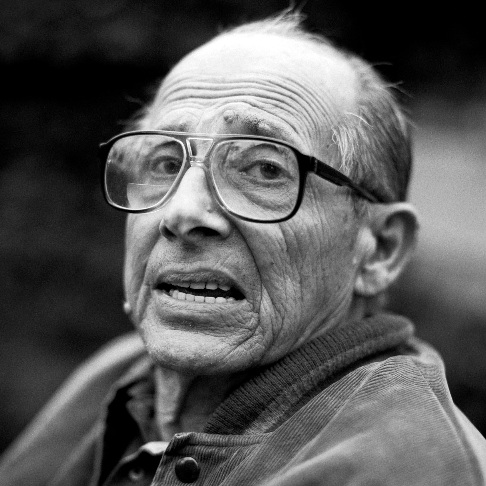 A close-up black and white photo of an elderly man wearing glasses, with visible wrinkles and gray hair, looking slightly to the side.