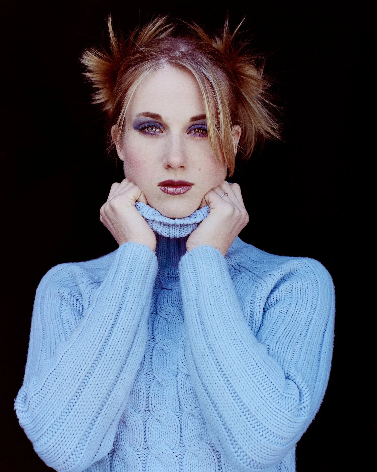 A woman with spiky reddish hair, wearing a light blue cable knit sweater, poses against a black background. She has blue eyeshadow, dark lipstick, and is holding the collar of her sweater with both hands.