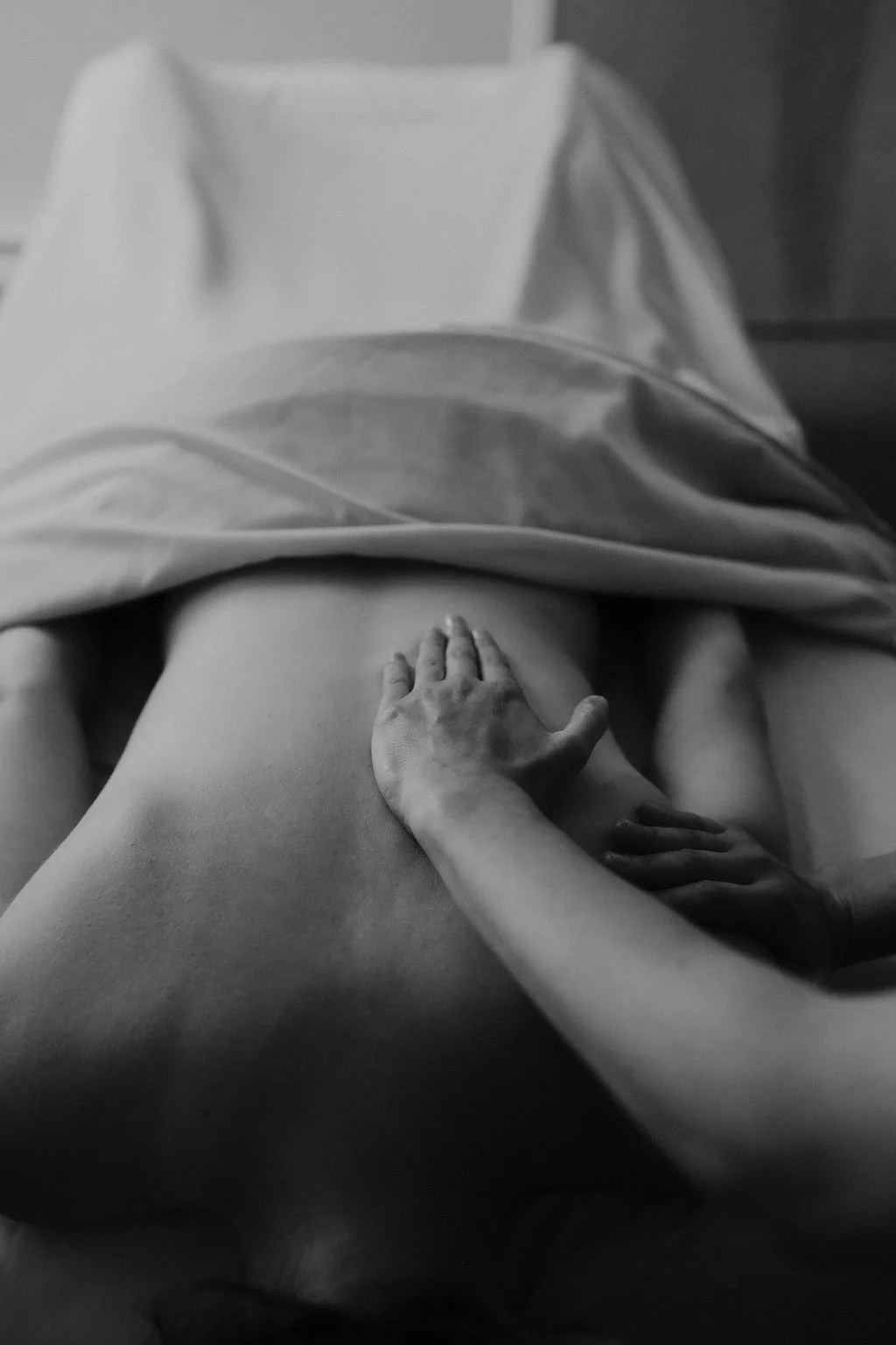 Black and white photo of a person lying on a bed, covered partially by a blanket, with one hand resting on their stomach and the other hand holding the person's arm.