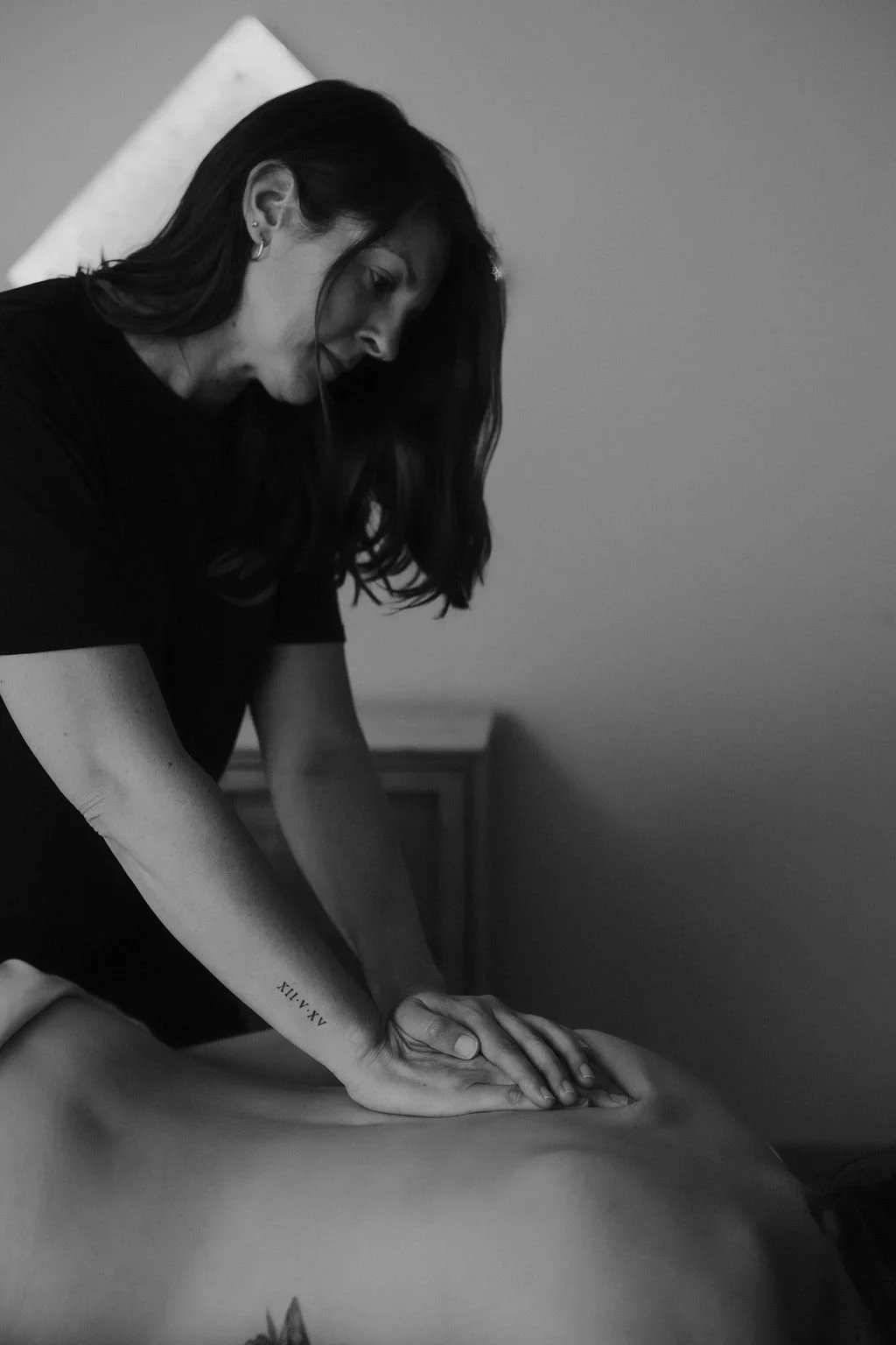 A woman giving a massage on a person's back, indoors with a plain wall and a piece of furniture in the background, in black and white.