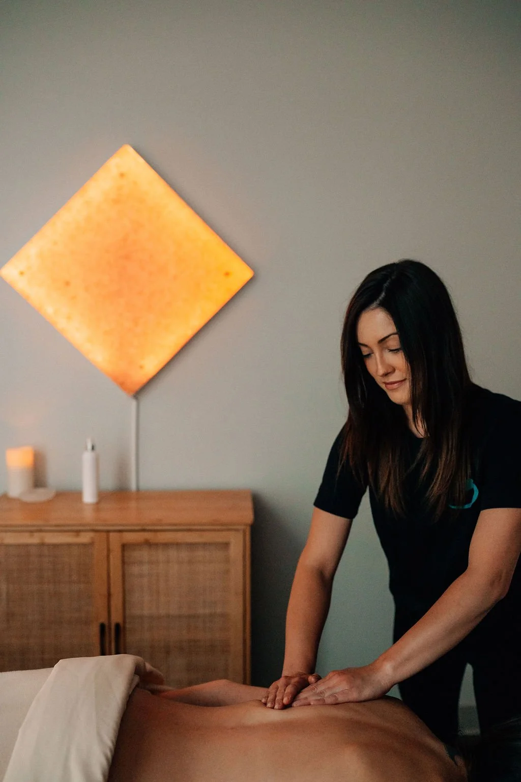 A woman giving a massage to a man lying face down on a massage table in a spa room, with candles and an orange wall hanging in the background.