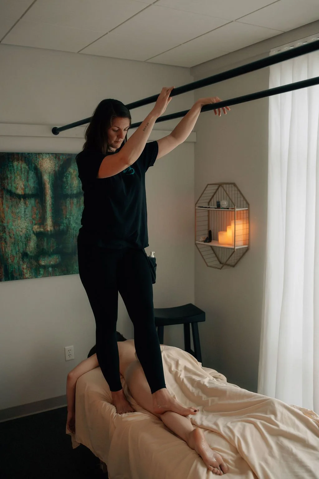 A woman standing on a bed with her arms raised holding a curtain rod, in a bedroom with a candle-lit shelf and a blanket-covered figure lying face down on the bed.