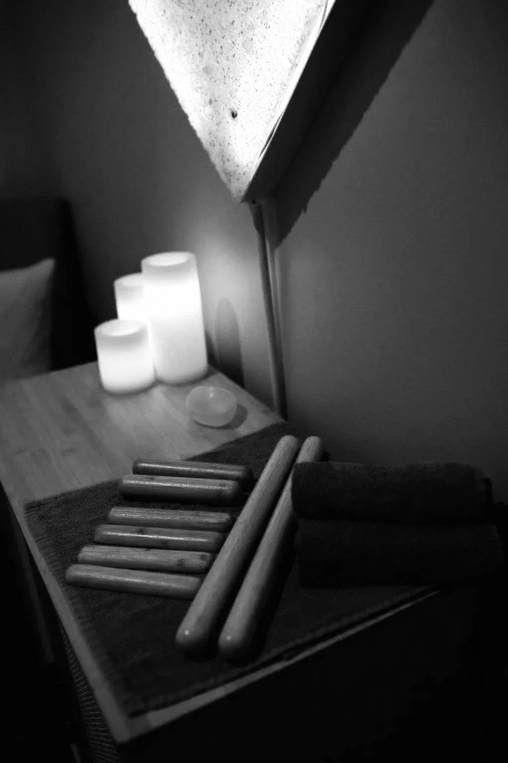 Black and white photo of wooden massage sticks on a tray, candles on a table, and a lamp on the wall in a dimly lit room.