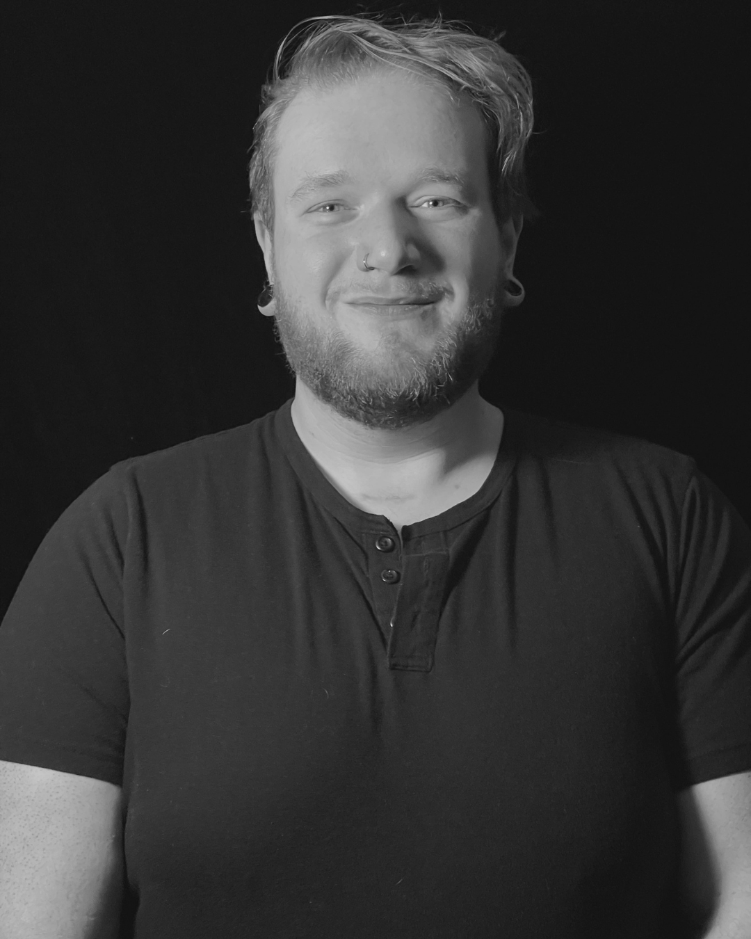 Black and white portrait of a smiling person with light-colored hair, beard, earrings, and nose ring, wearing a dark shirt, against a dark background.