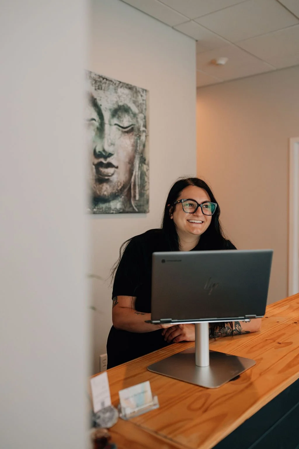 A woman with glasses and long dark hair smiling at a computer monitor on a wooden counter in a room with beige walls and a Buddha painting on the wall behind her.
