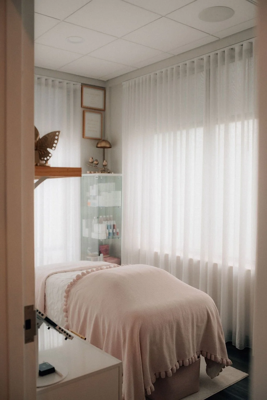 A cozy bedroom with a bed covered in a beige blanket, white sheer curtains on the windows, and decorative frames and objects on the wall and shelves.