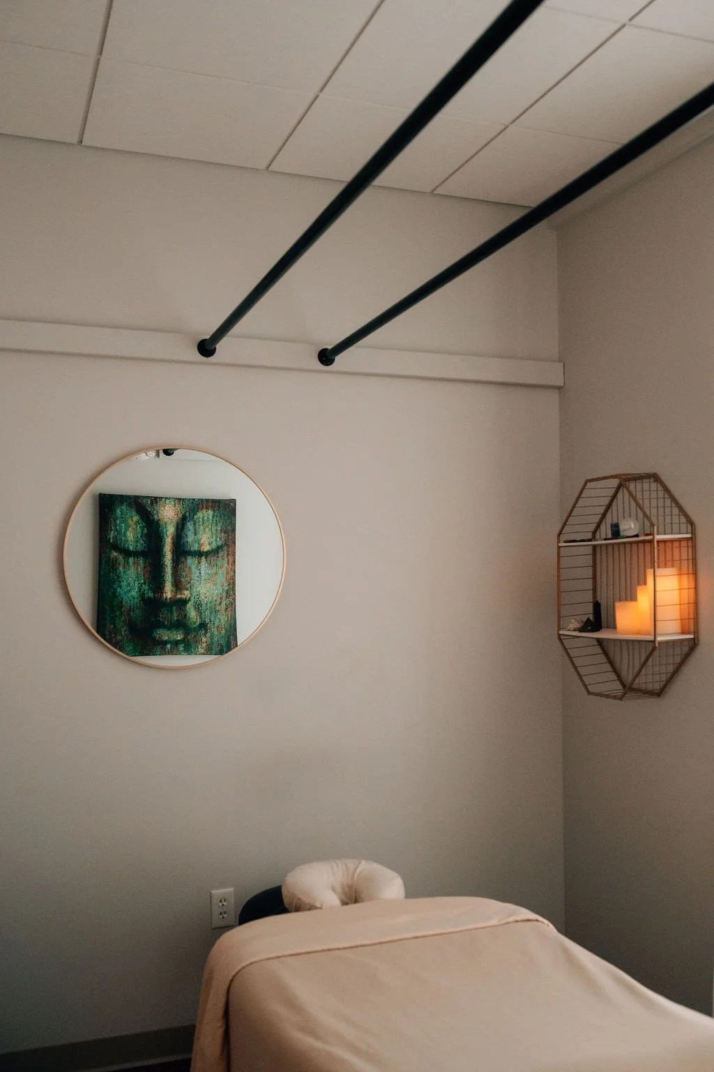 A room with a wall-mounted art piece of a green Buddha face, a circular mirror, a wooden hexagonal shelf with candles and decorative items, a massage table with a beige sheet and a rolled towel, and ceiling pipes overhead.