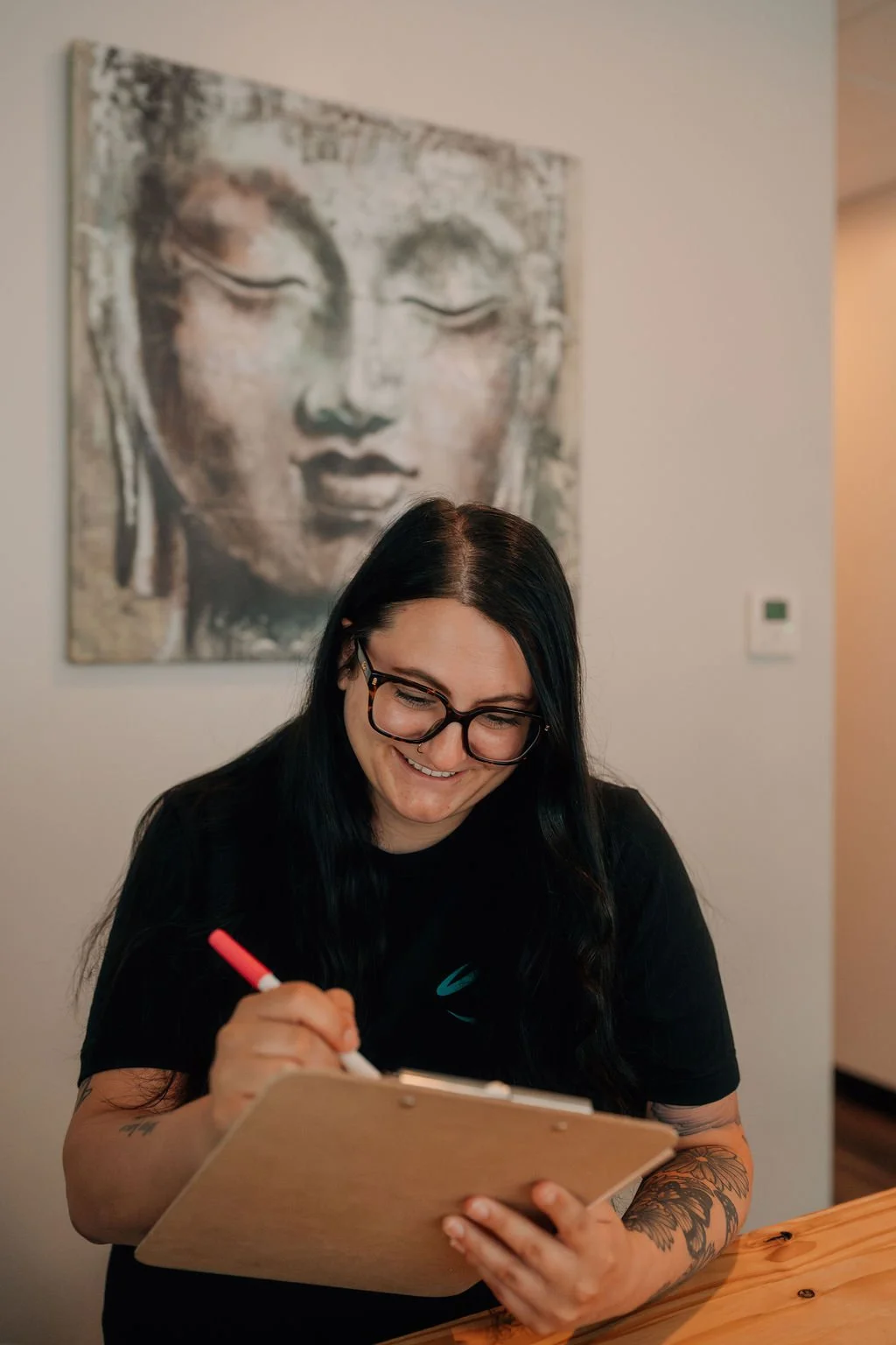 A woman with black hair and glasses smiling while writing on a clipboard, sitting at a wooden table, with a large Buddha painting hanging on the wall behind her.