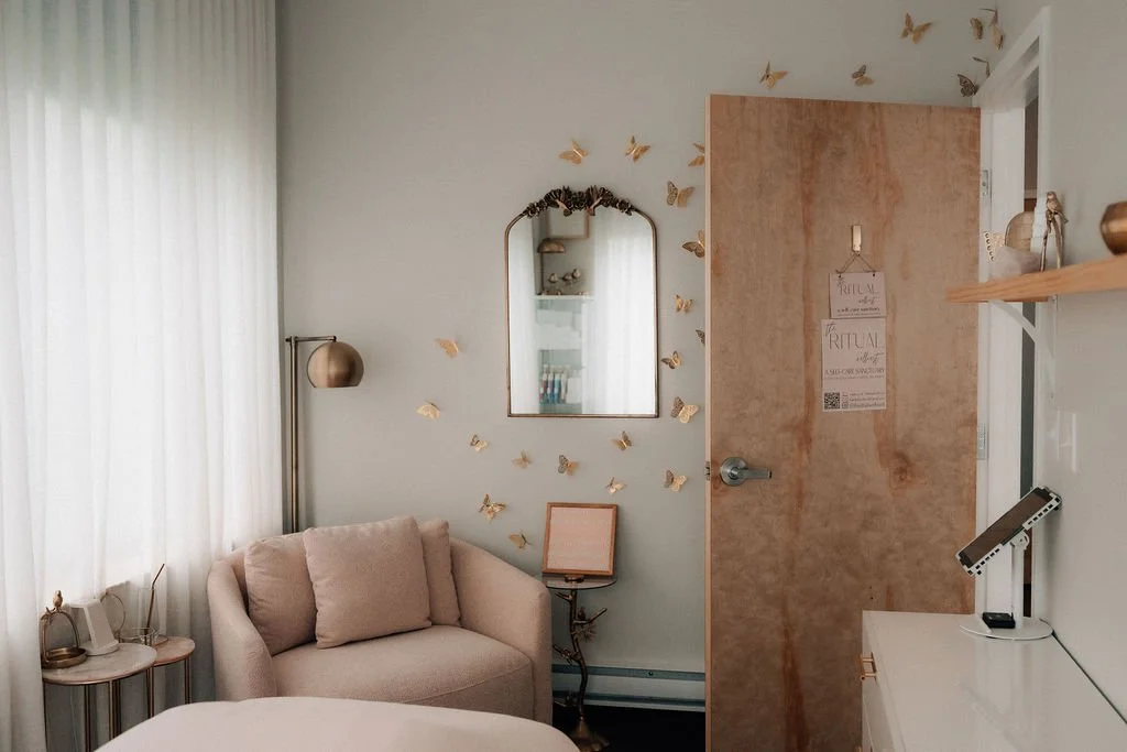 A cozy bedroom corner with a beige armchair, a small side table, gold butterfly wall decals around a mirror, and a wooden door.